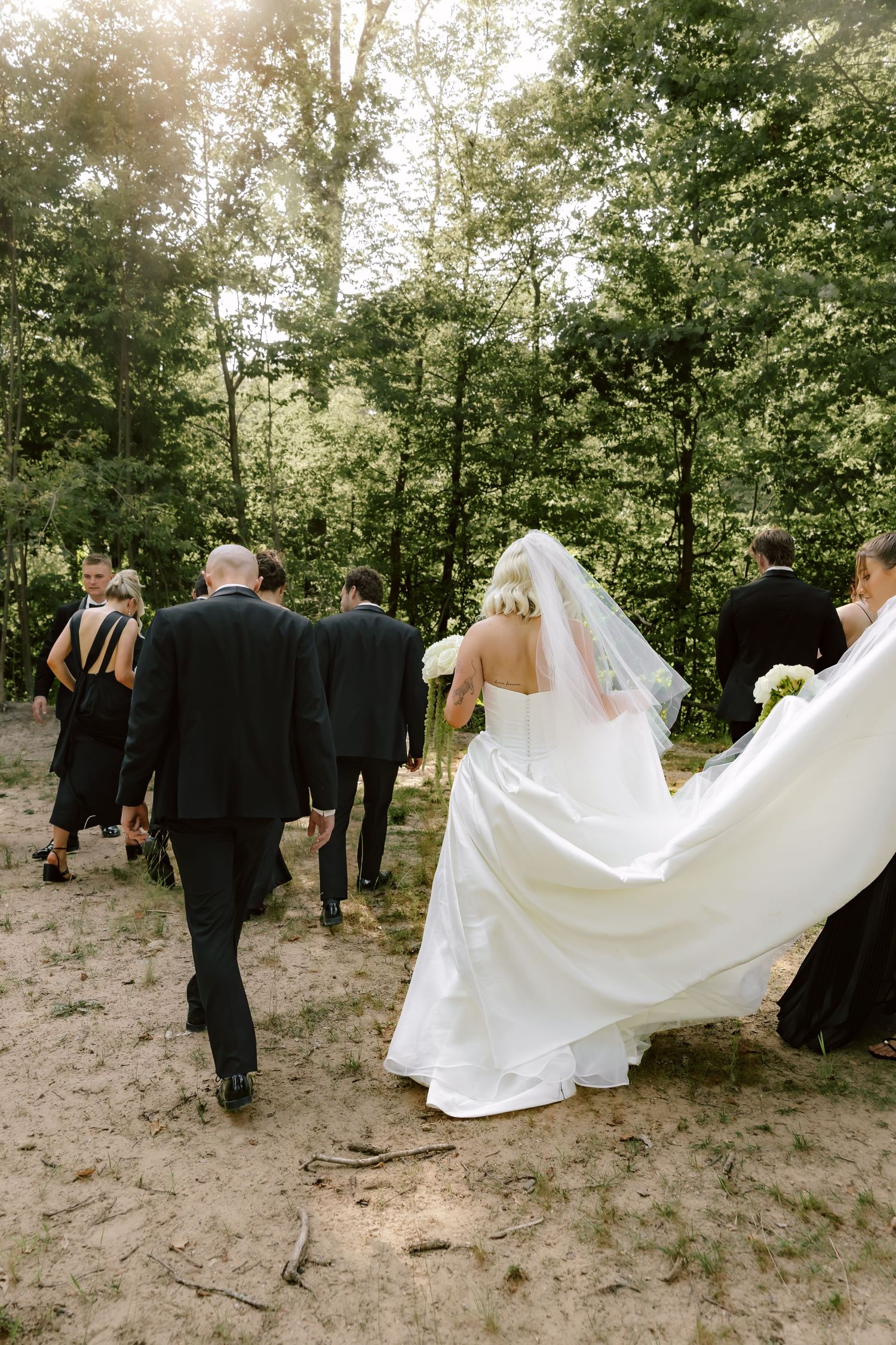 Wedding party walks through wooded area with flowing white wedding dress trailing behind.