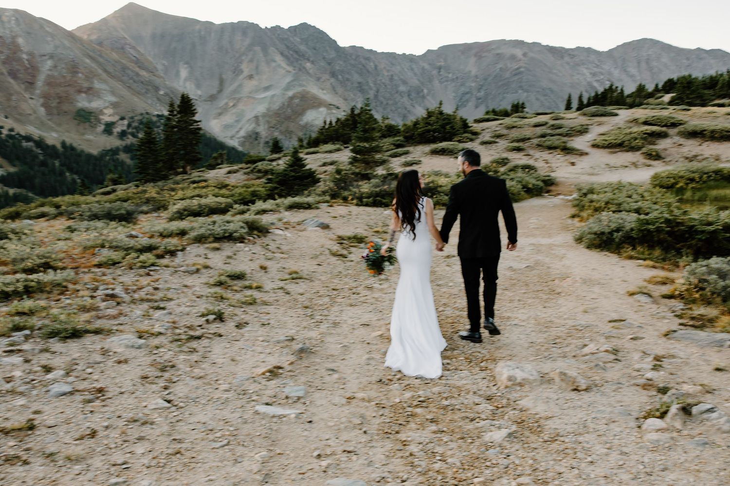 A couple in formal attire walks hand in hand along a mountain trail with rugged peaks in the background.
