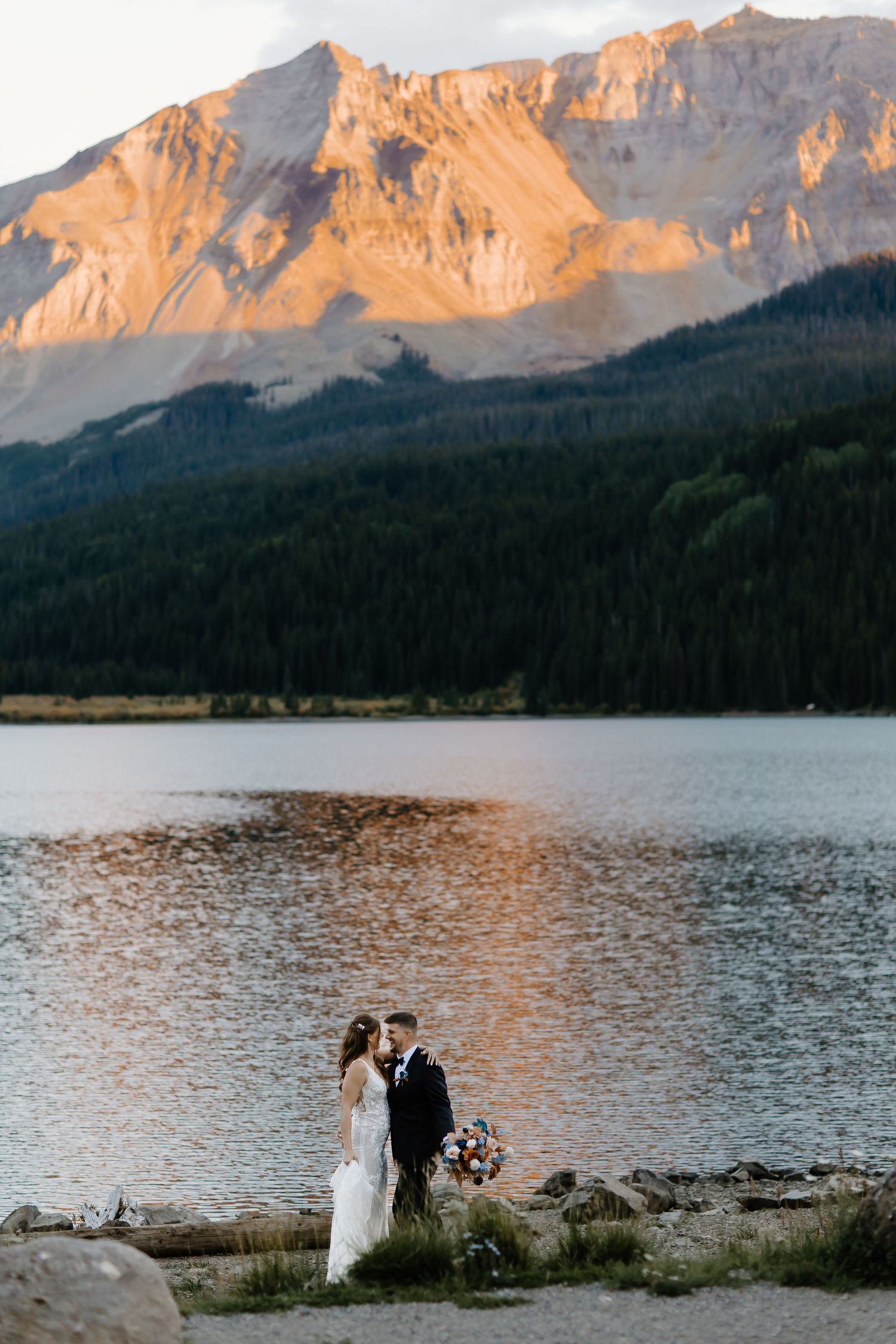 Wedding couple stands by alpine lake with dramatic mountain peaks glowing at sunset.