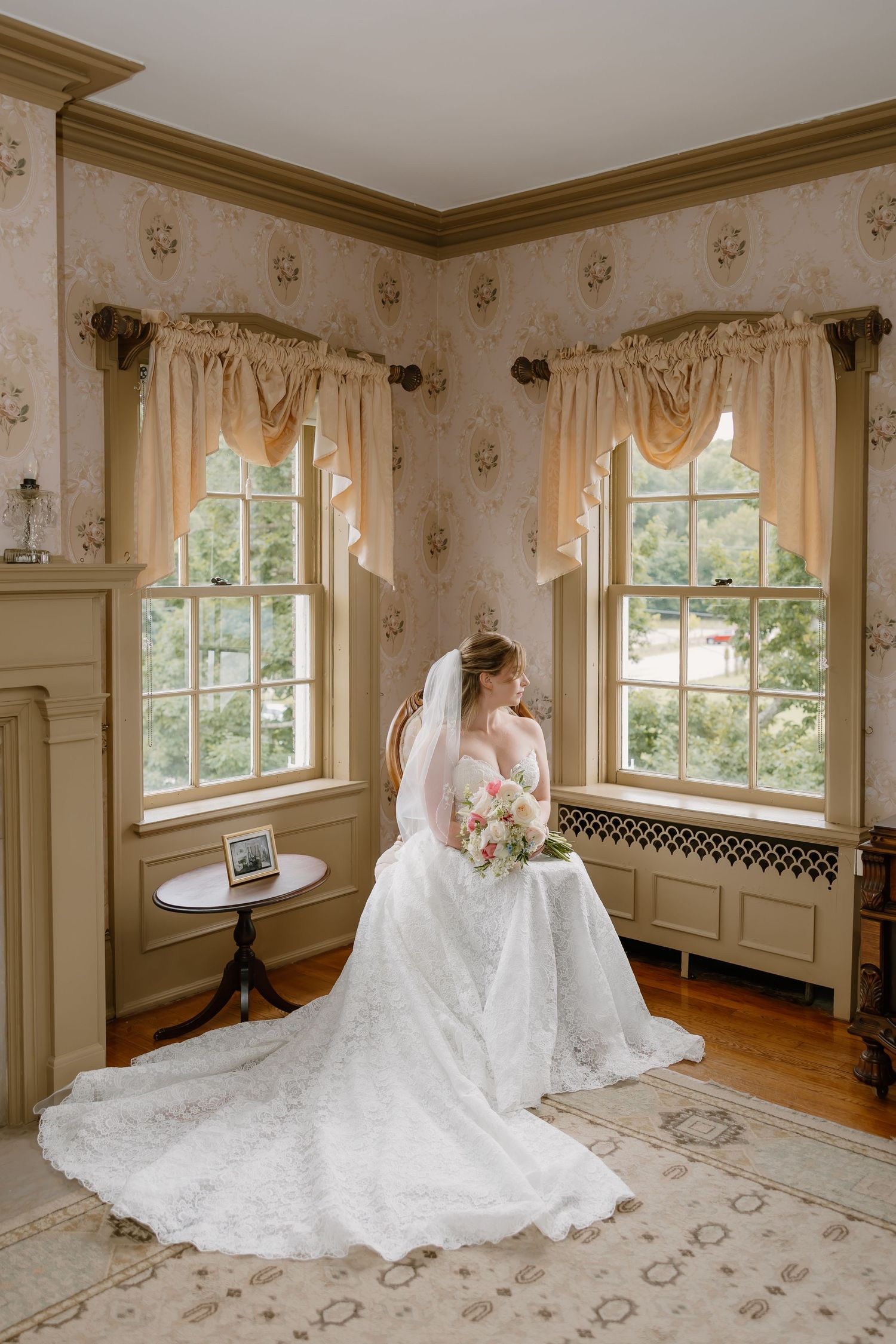 Bride in lace wedding gown and veil sits by vintage windows holding bouquet in elegant floral wallpapered room.