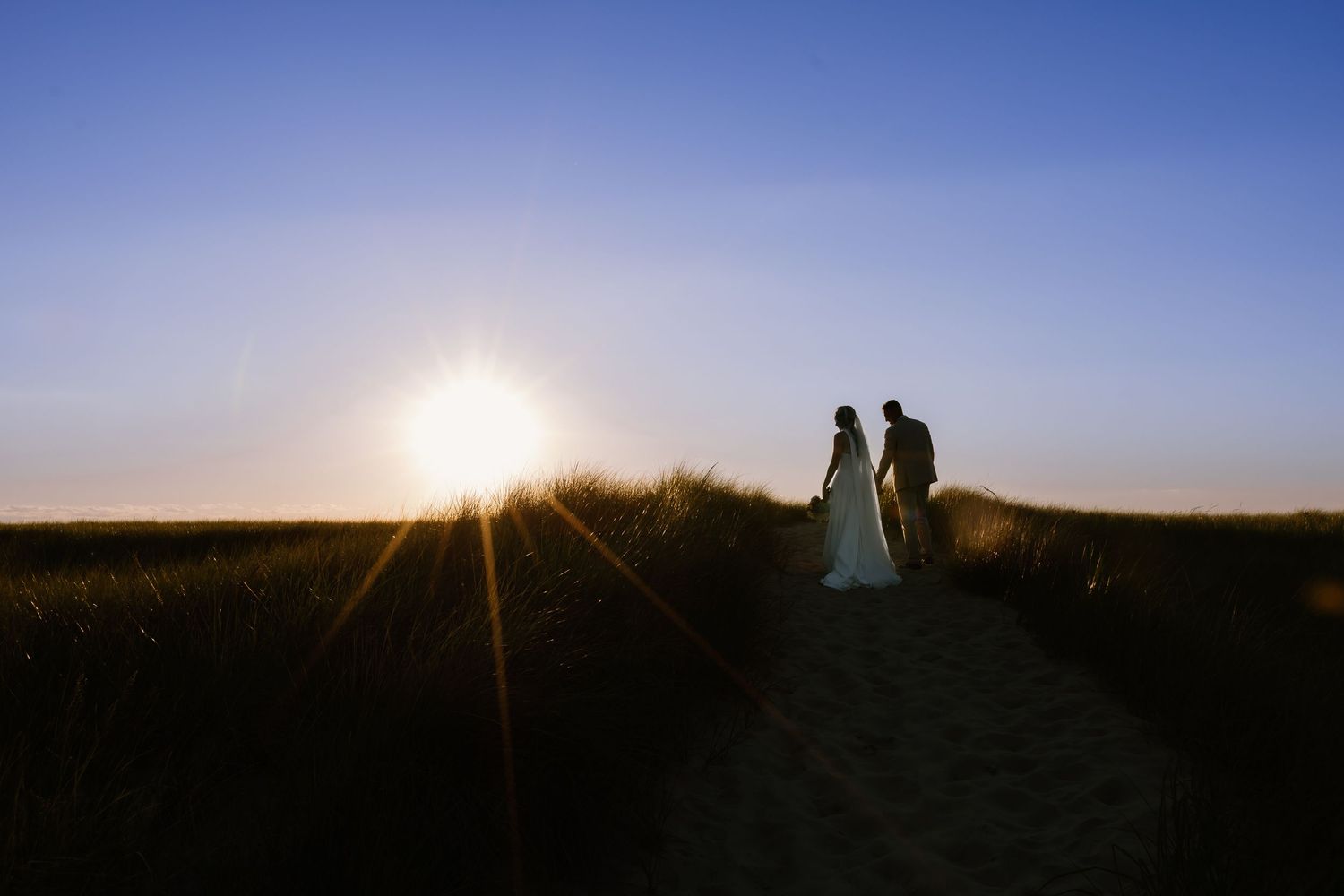 Silhouette of bride and groom holding hands on sandy path at golden sunset with sun flare and blue sky.