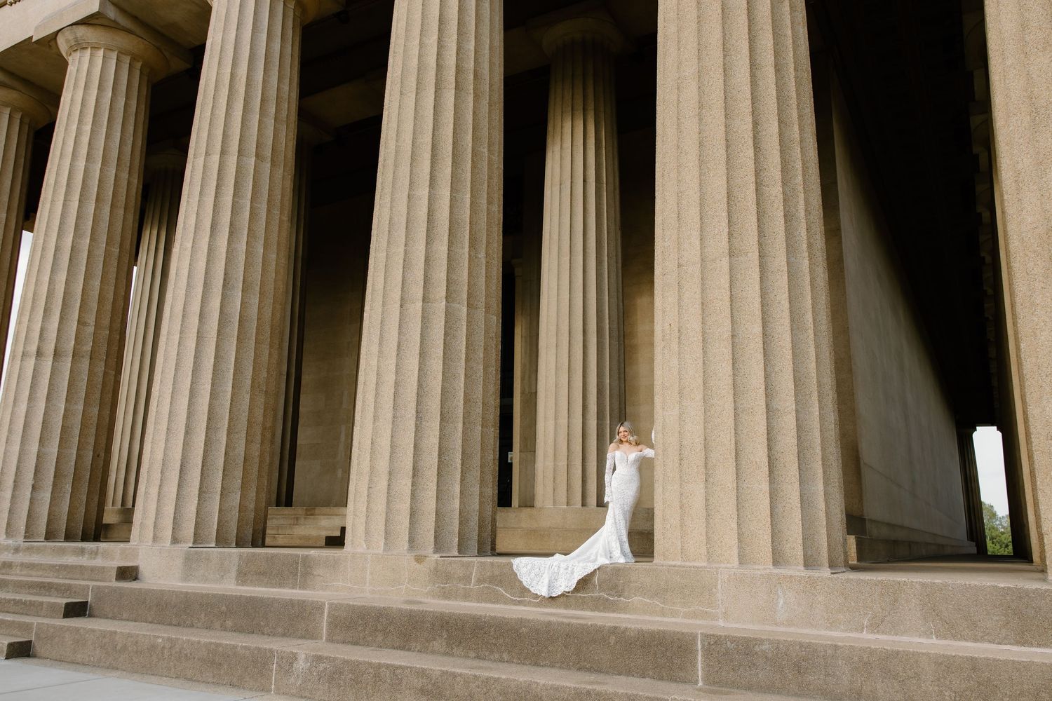 Bride in white gown poses on steps of grand columned building resembling a Greek temple.