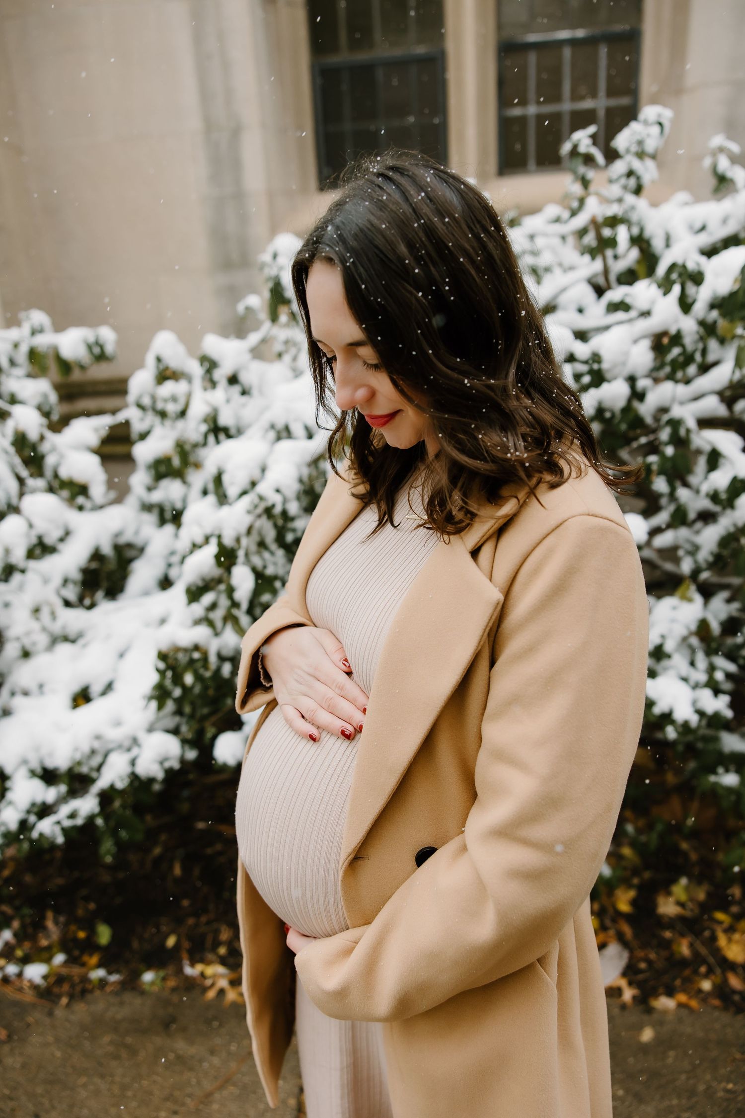 Winter maternity photo featuring a cream knit dress and camel coat against snowy backdrop.