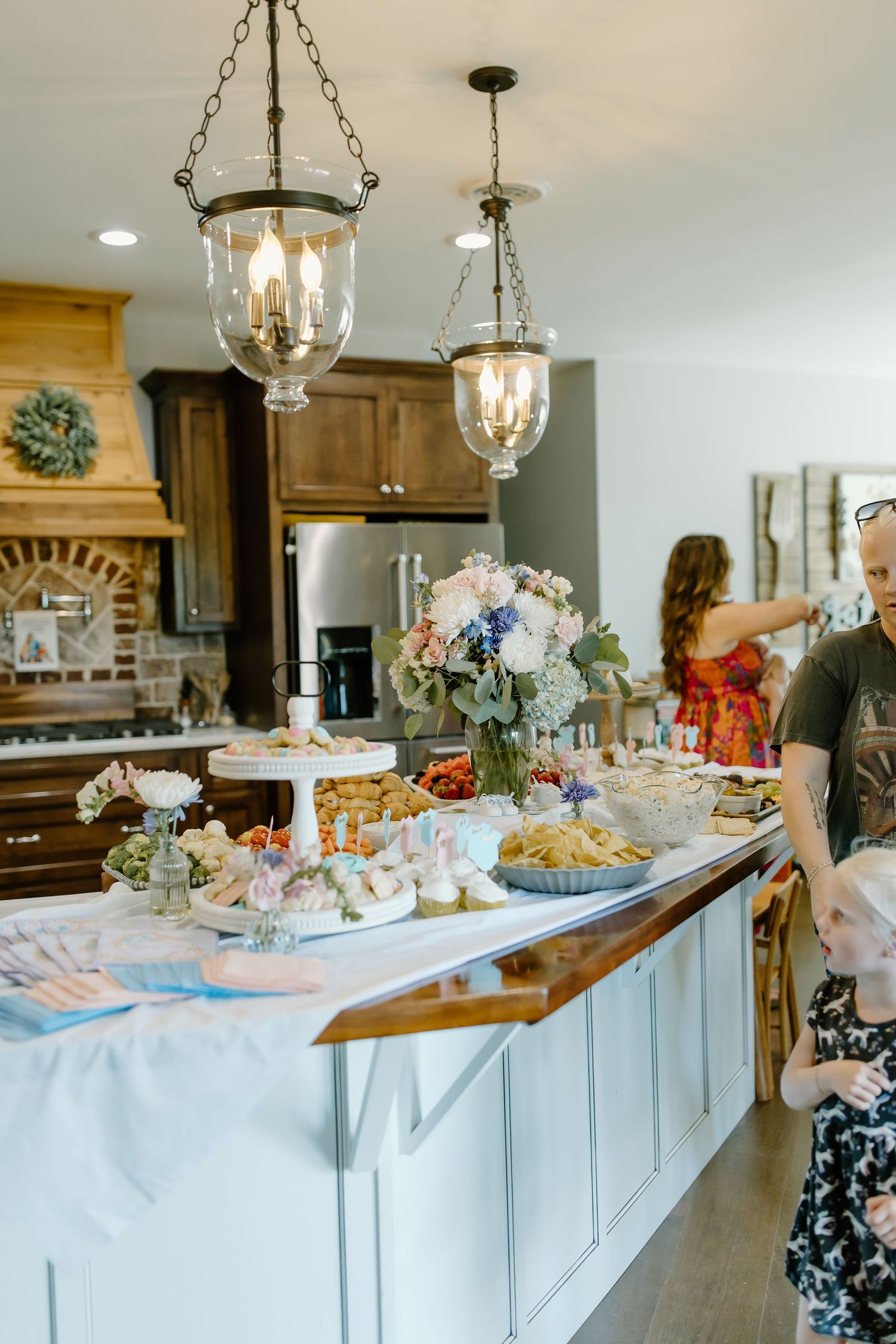 Elegant kitchen island decorated with floral arrangements and party food displays under glass pendant lighting.