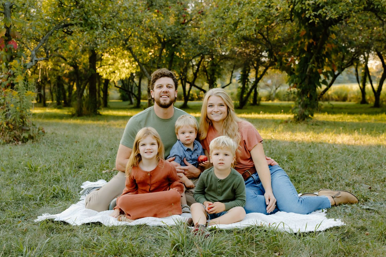A family relaxes together on a white blanket during an autumn photo session in a park setting.
