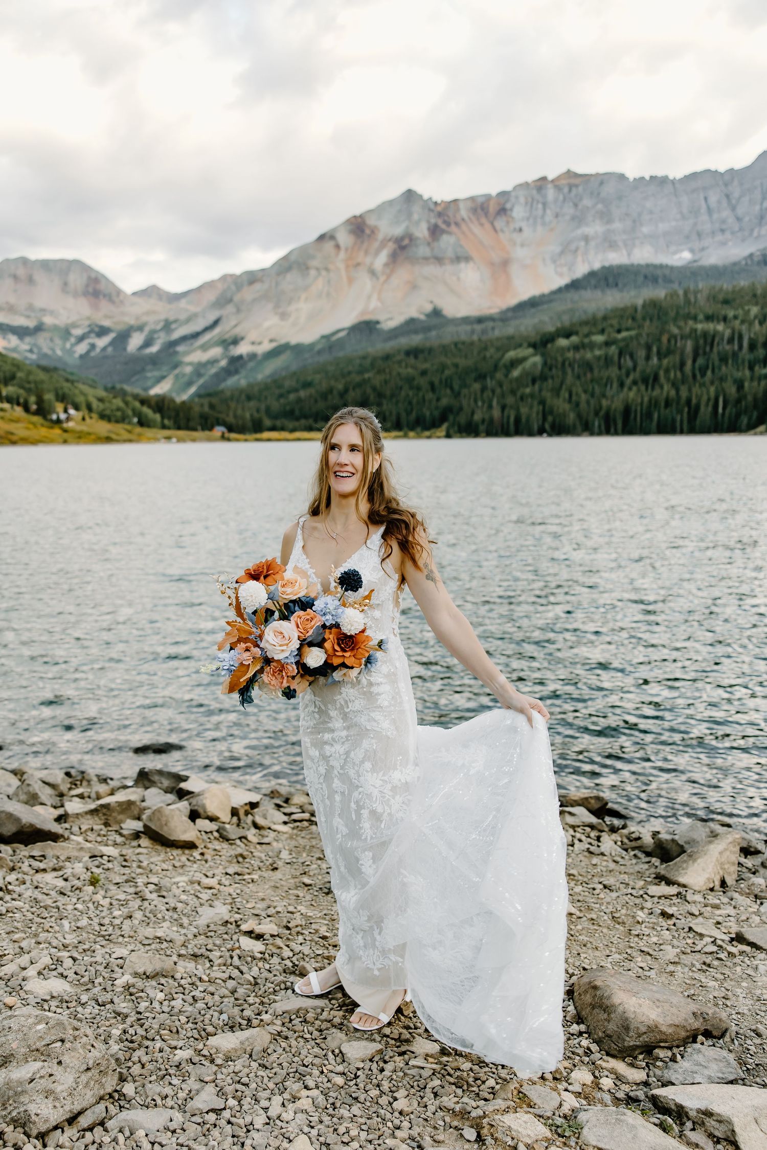 White lace wedding dress photographed against mountain lake backdrop with orange bouquet.