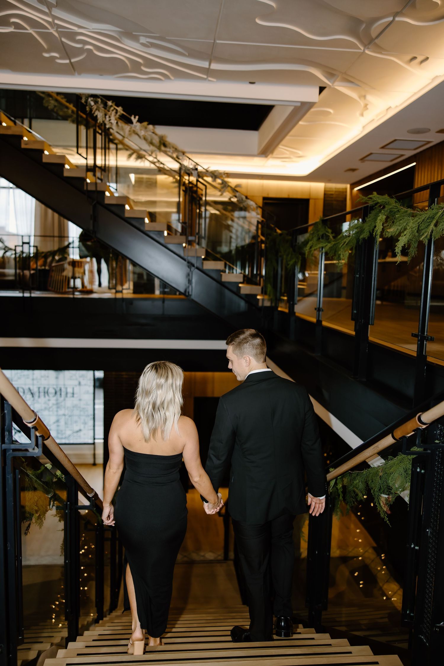 Couple ascending elegant staircase in formal attire at upscale venue with decorative lighting.