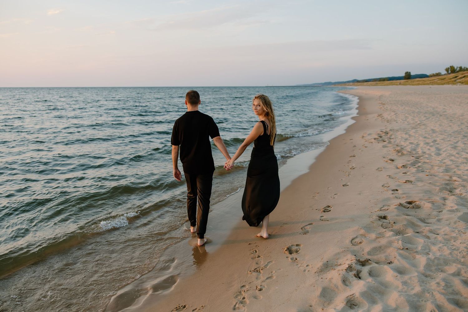 Couple walking along shoreline holding hands during sunset.