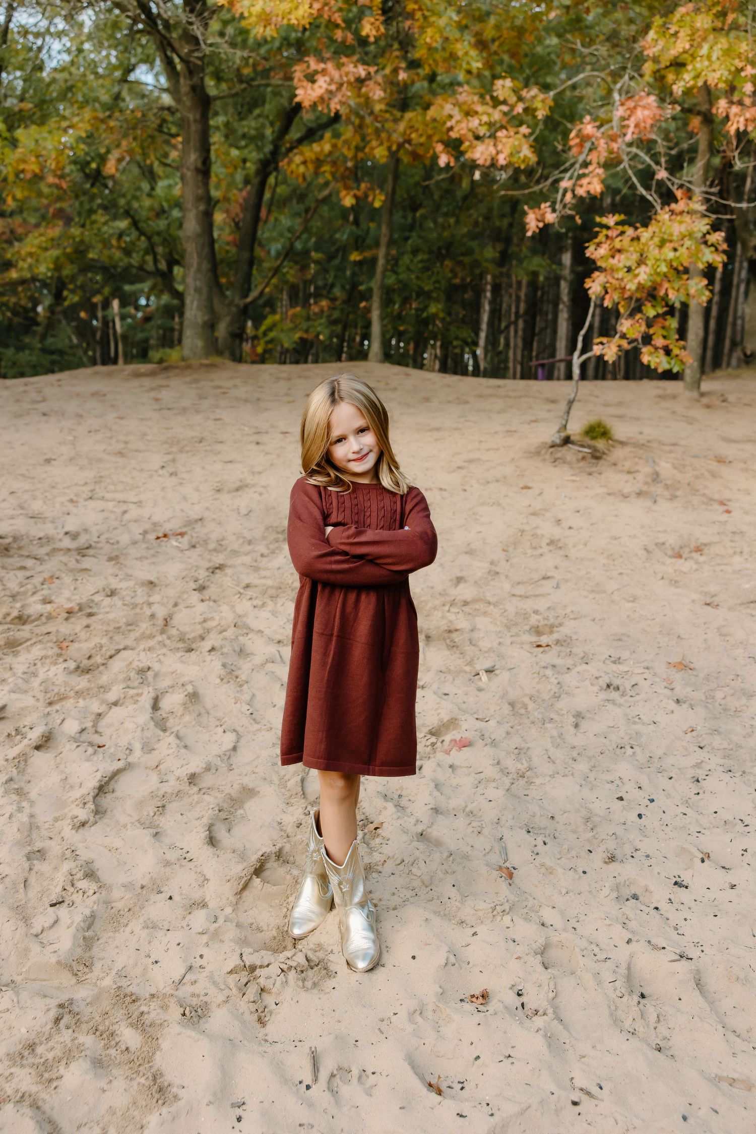 Child in burgundy dress stands alone on sandy ground with autumn trees in background.