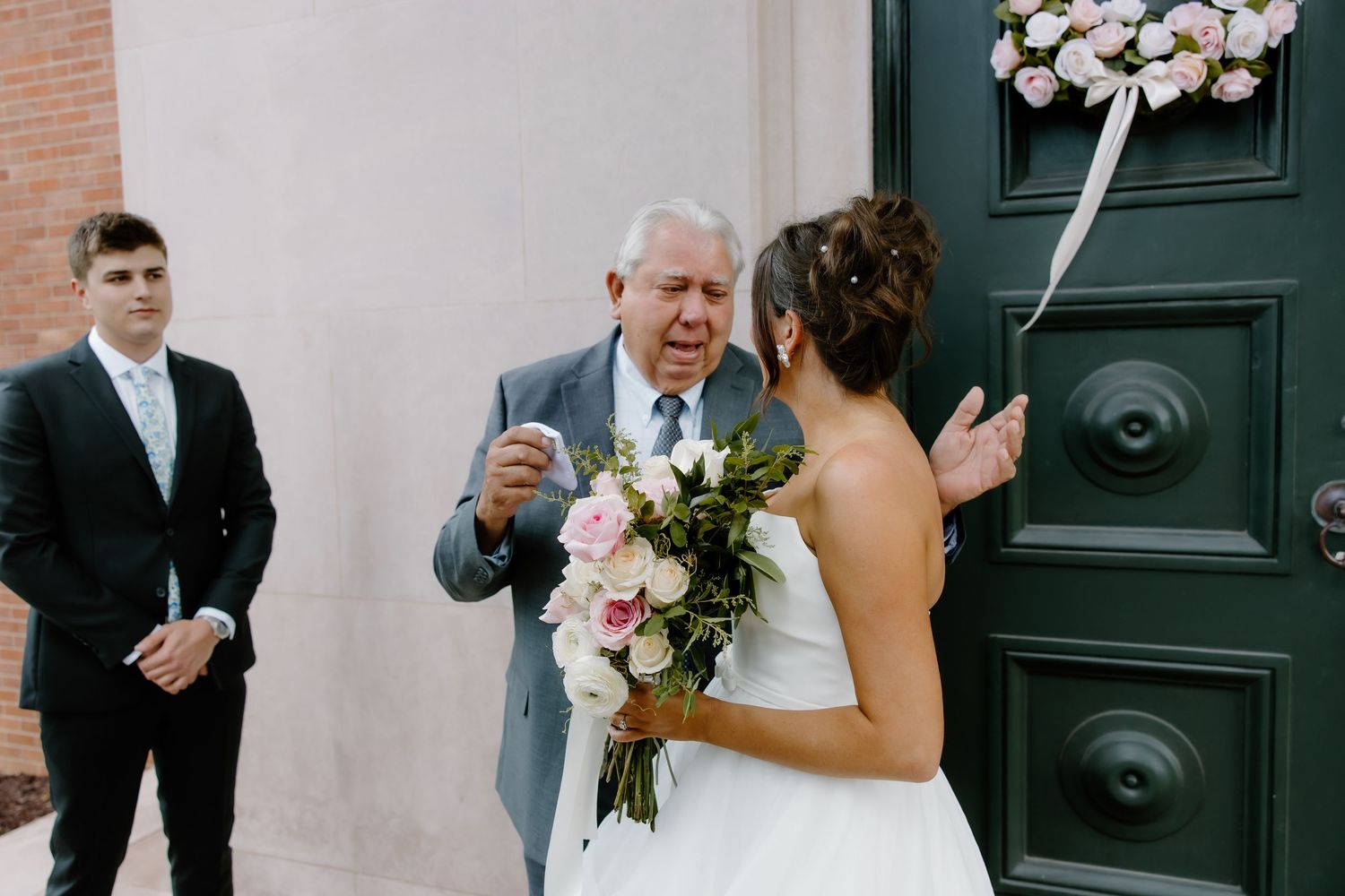 A bride in a white gown holds a pink bouquet while standing near a green door with a wreath.