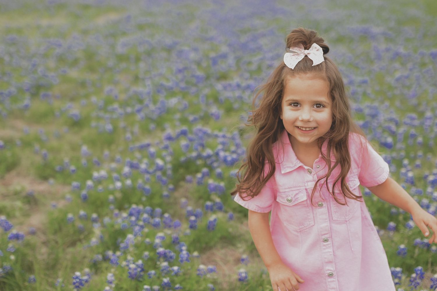 A smiling young girl in a pink dress with a bow in her hair stands in a field of purple bluebonnets.