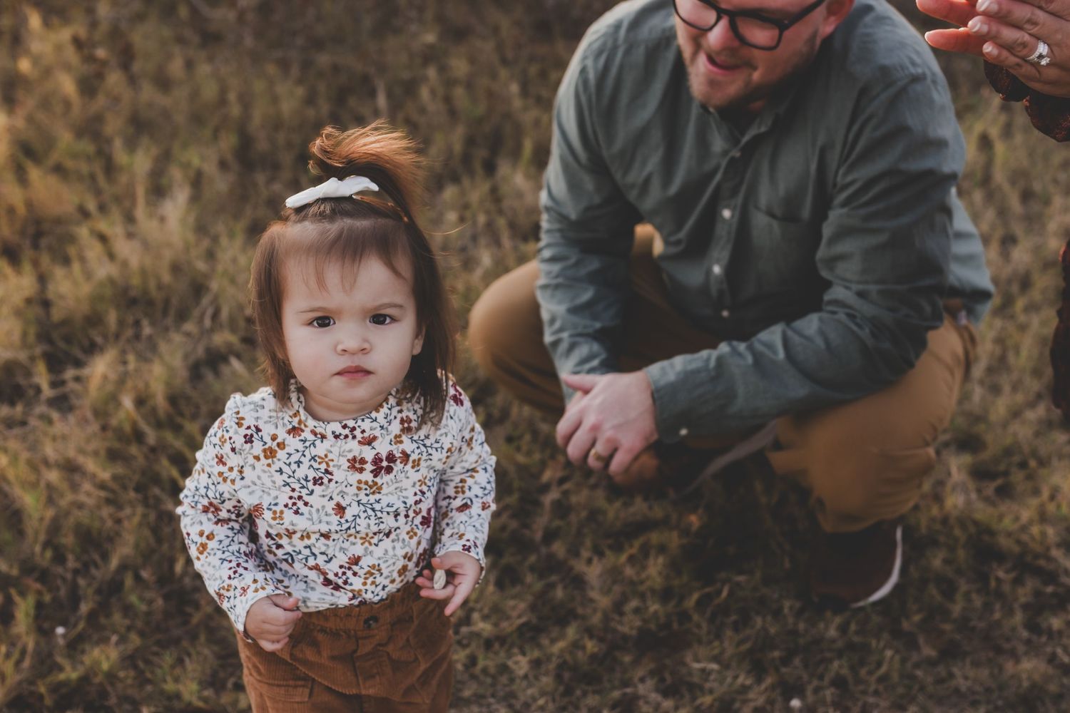 Toddler girl in floral top and brown skirt stands in field while father crouches behind her during fall family photo session.