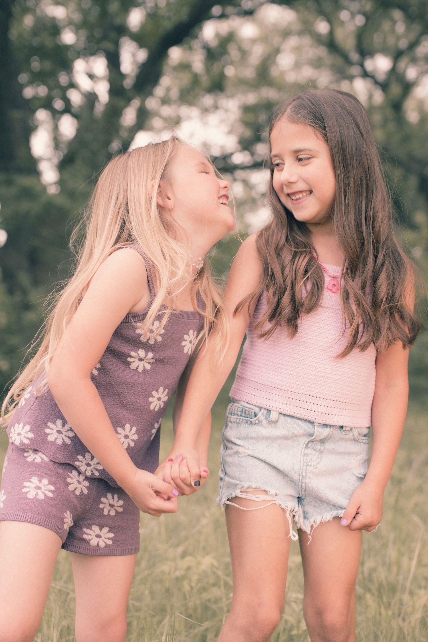 Two young girls holding hands and laughing together outdoors in a green field, wearing summer outfits.