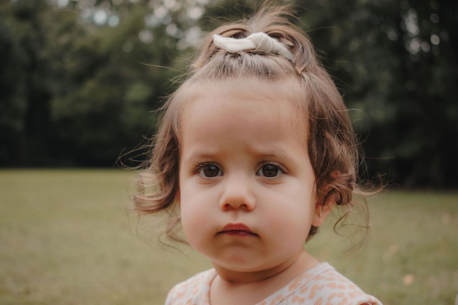 A close-up portrait of a toddler girl with a white bow outdoors in a park.