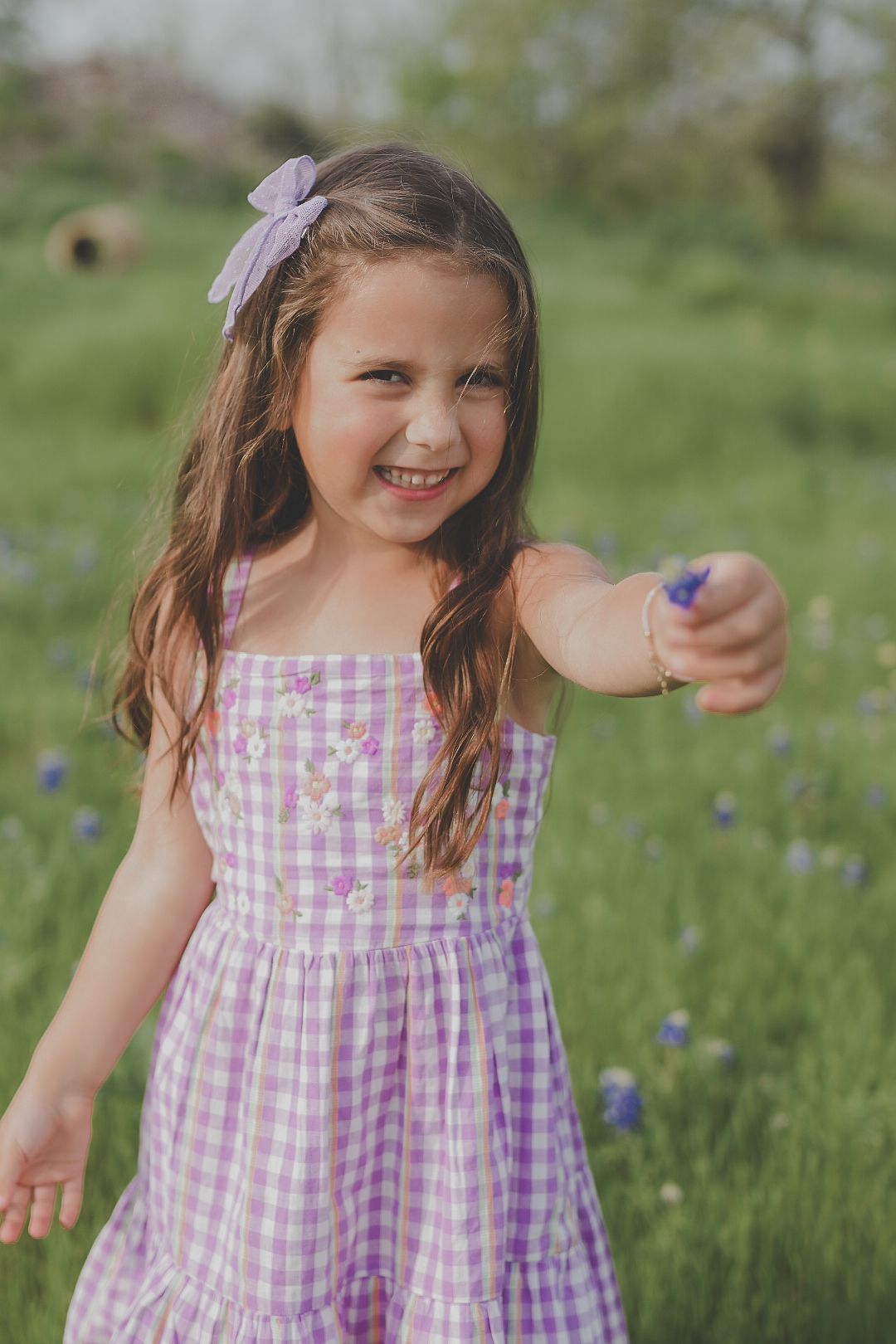 Cheerful young girl in purple gingham dress holds out a bluebonnet flower in a spring wildflower field portrait.