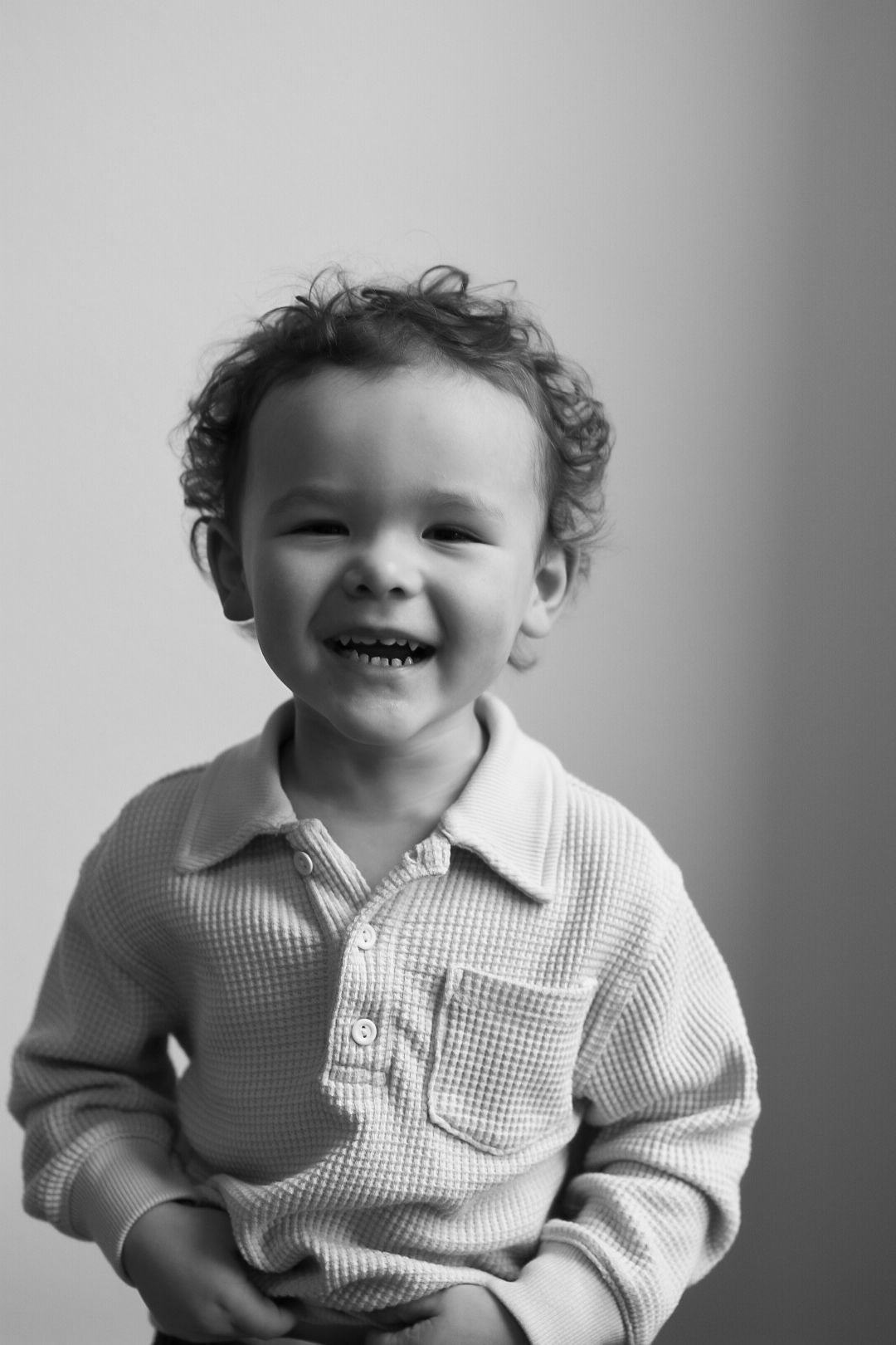 A curly-haired toddler boy smiles brightly in a black and white portrait wearing a polo shirt.