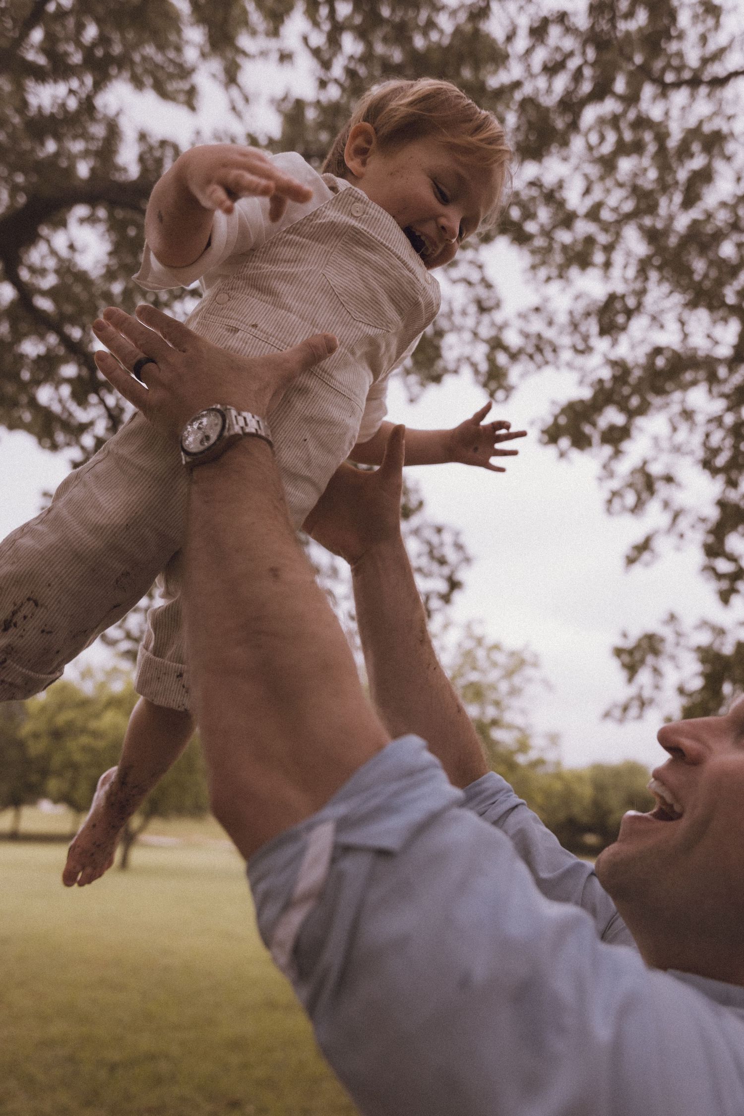 Father joyfully lifting toddler high in the air outdoors in a lush green park under a large tree.