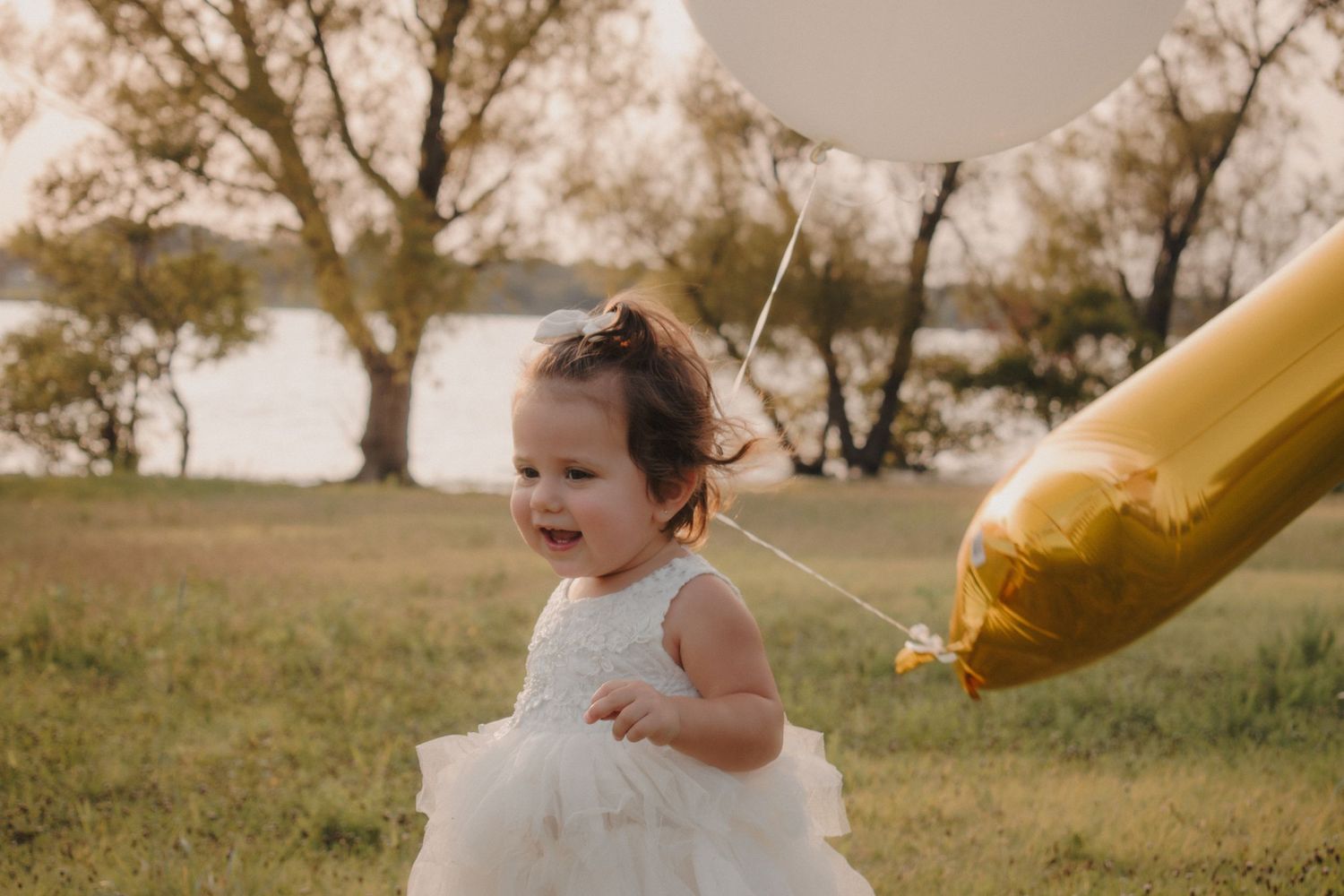 A joyful toddler in a white dress runs through a park holding gold and white balloons.