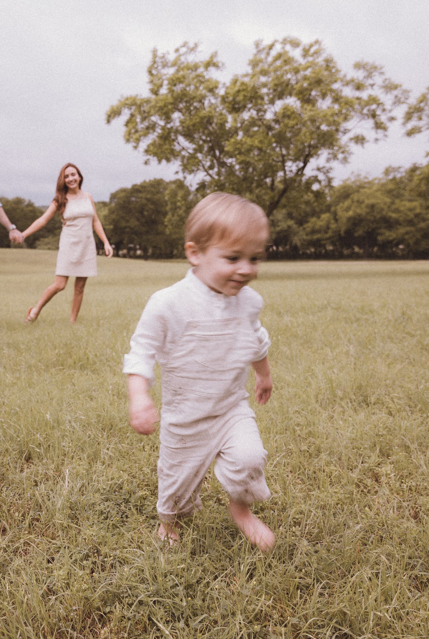 Happy toddler running barefoot in a green park with smiling mother in background on cloudy day.
