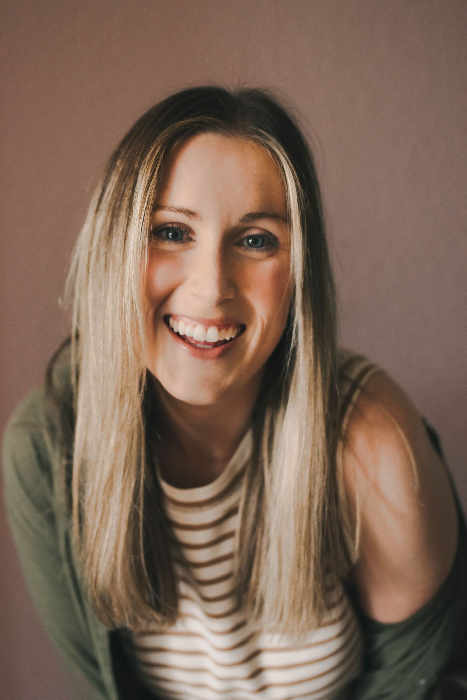 Smiling blonde woman with blue eyes wearing a striped shirt and green cardigan against a pink wall.