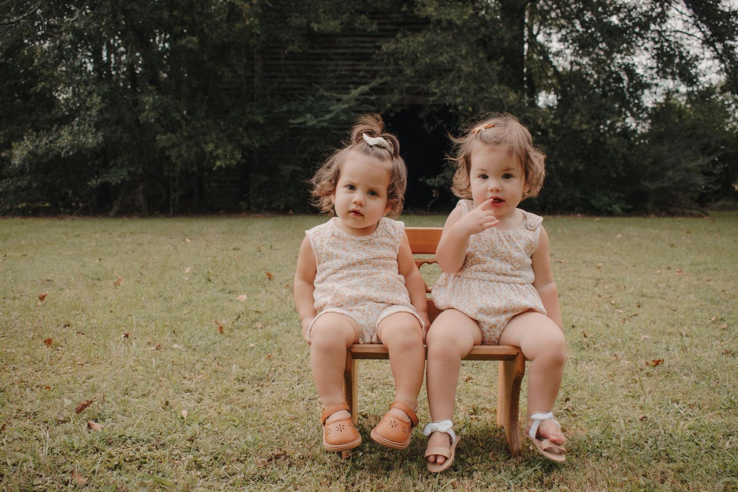 Two baby girls in matching cream rompers sit together on a small wooden bench outdoors.
