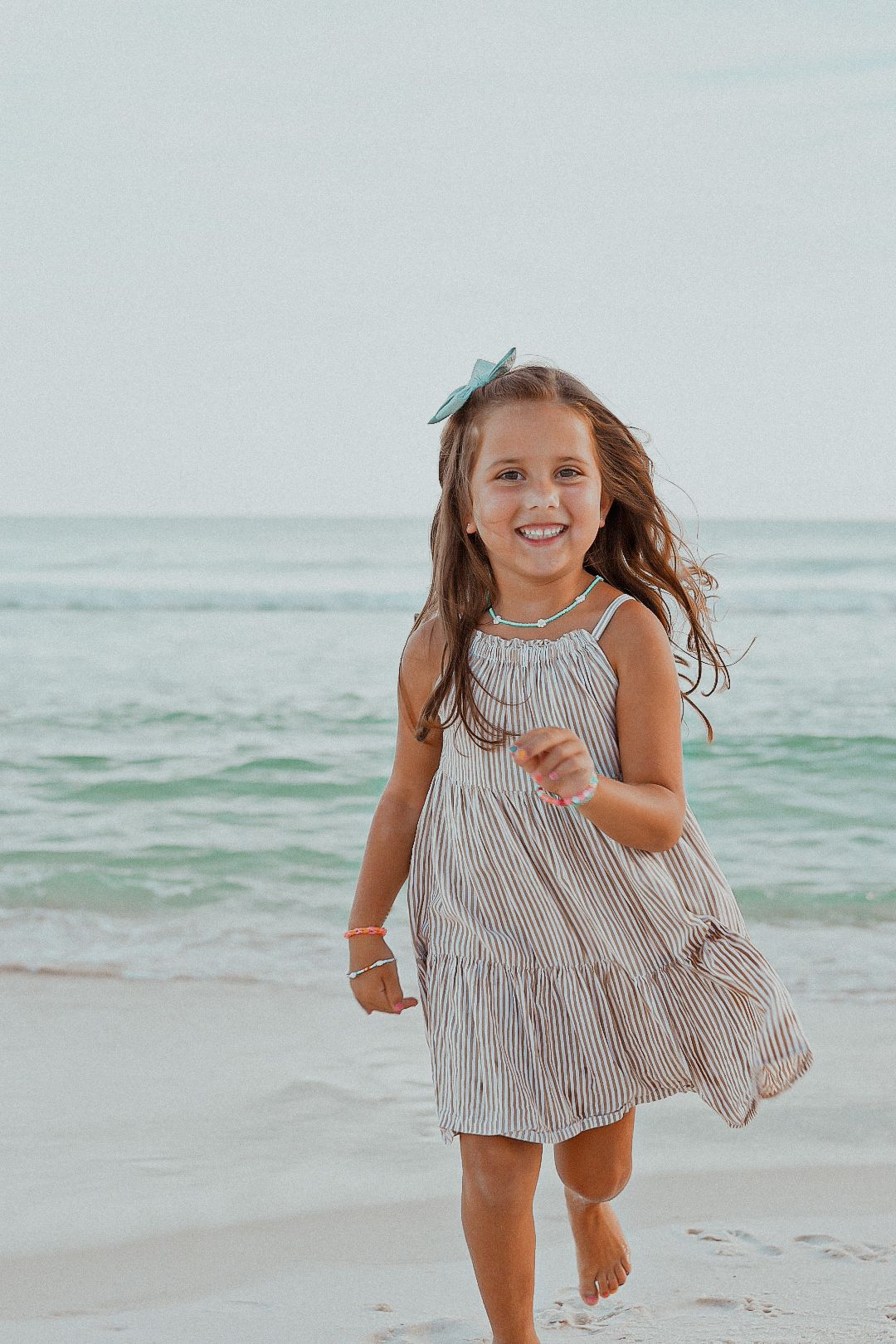 Happy young girl in a striped sundress running along a sandy beach with ocean waves behind her.