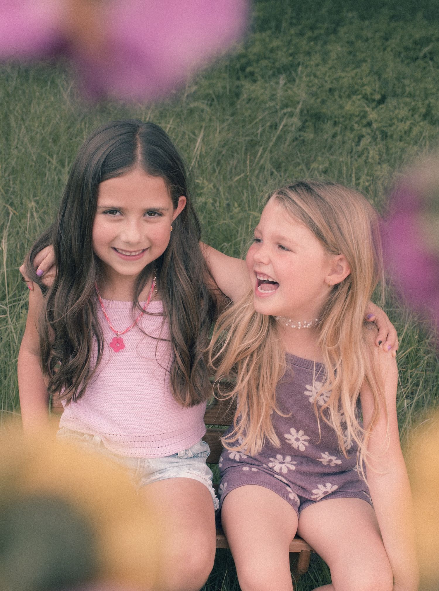 Two smiling girls sitting together outdoors in a field, wearing summer tops and floral accessories.
