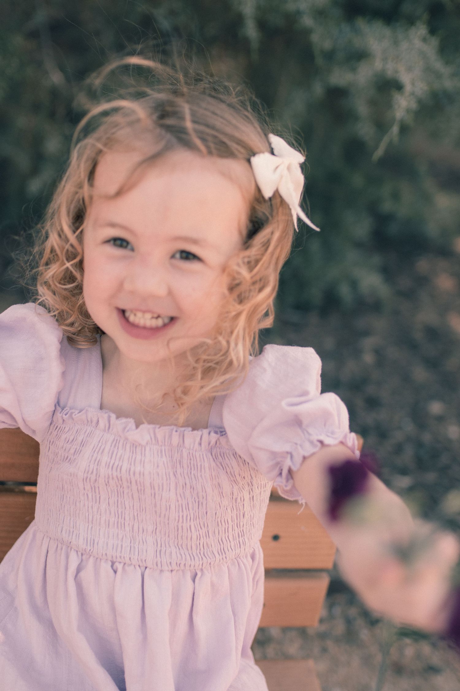 Smiling toddler girl with curly blonde hair and bow wearing a pink smocked dress, seated outdoors near green bushes.