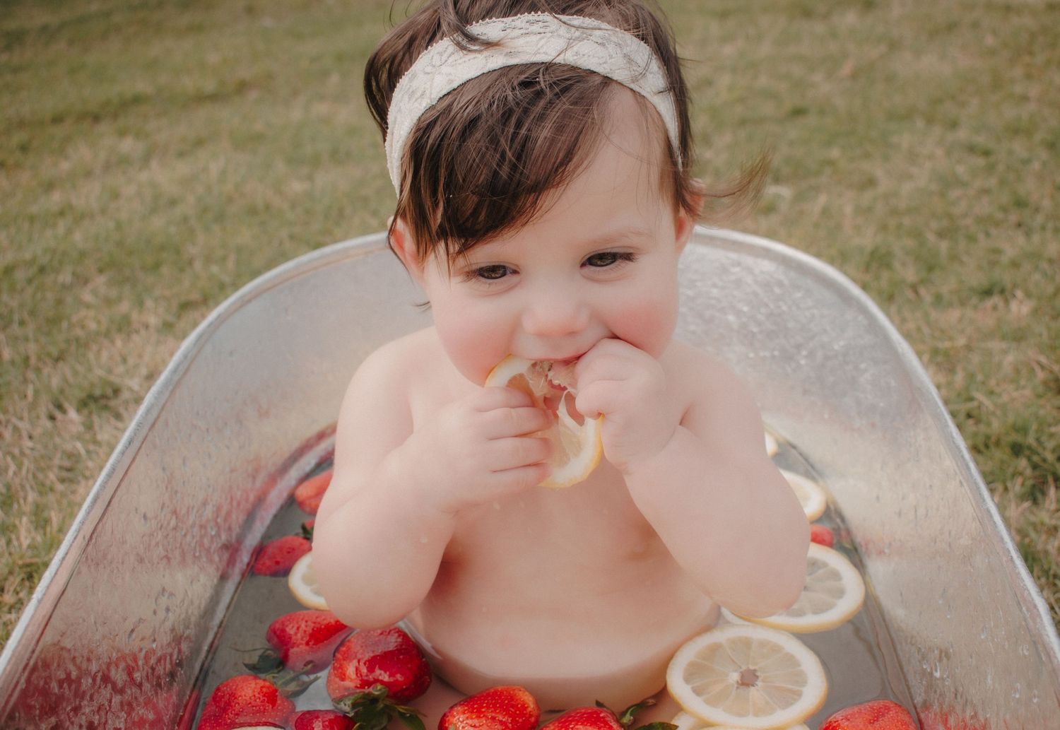 A cute baby girl sits in a metal tub with strawberries and lemons, nibbling a slice.