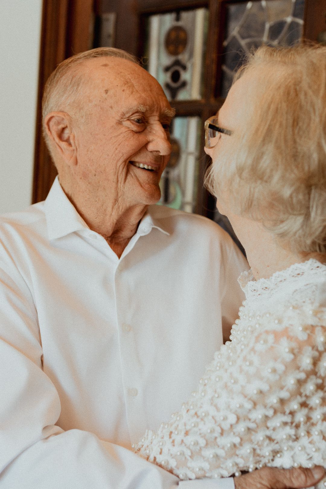 An elderly couple smiles warmly at each other, the man in a white shirt and woman in lace.