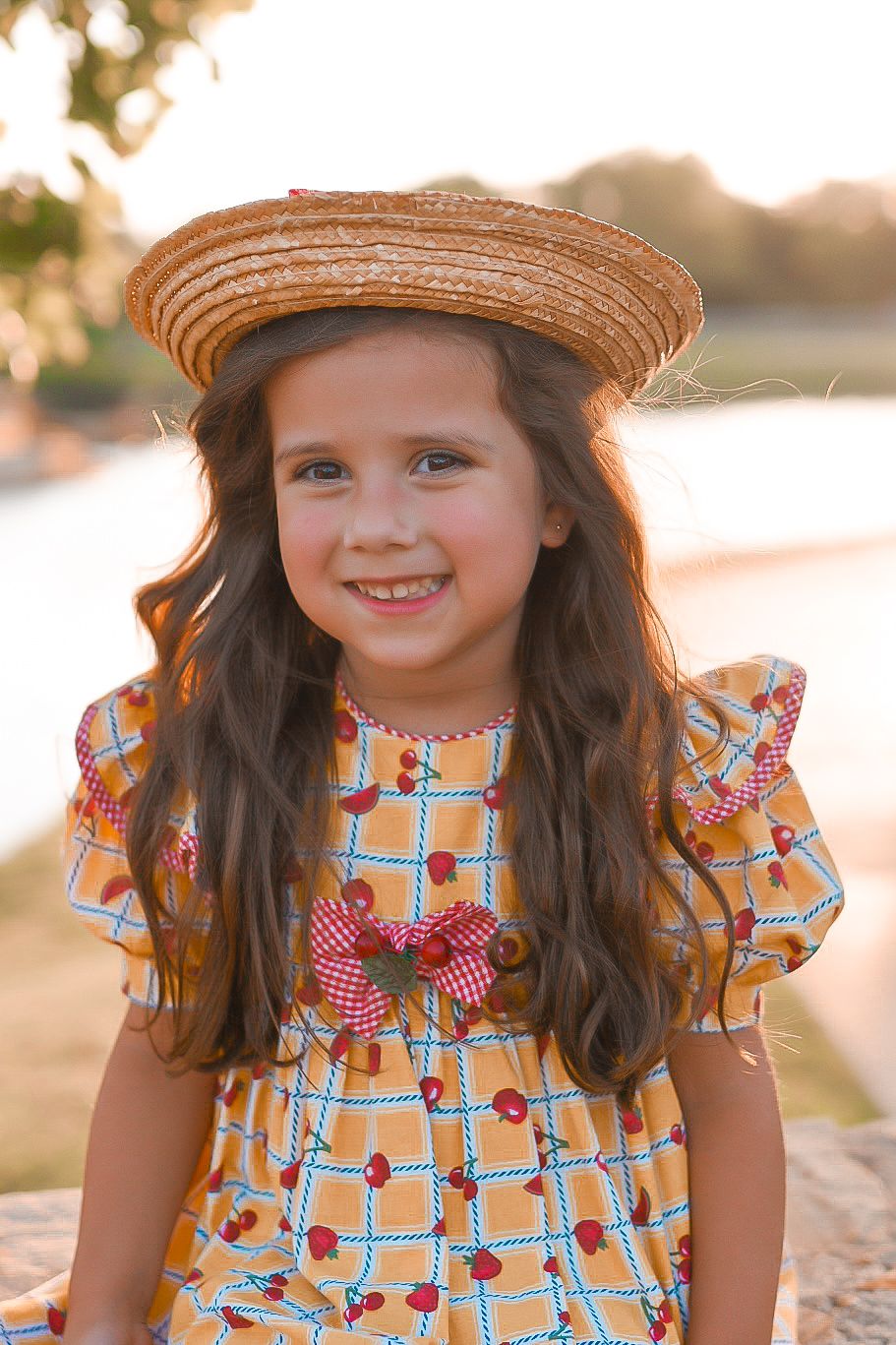 Smiling young girl in cherry print dress and straw hat poses outdoors near river during golden hour portrait session.