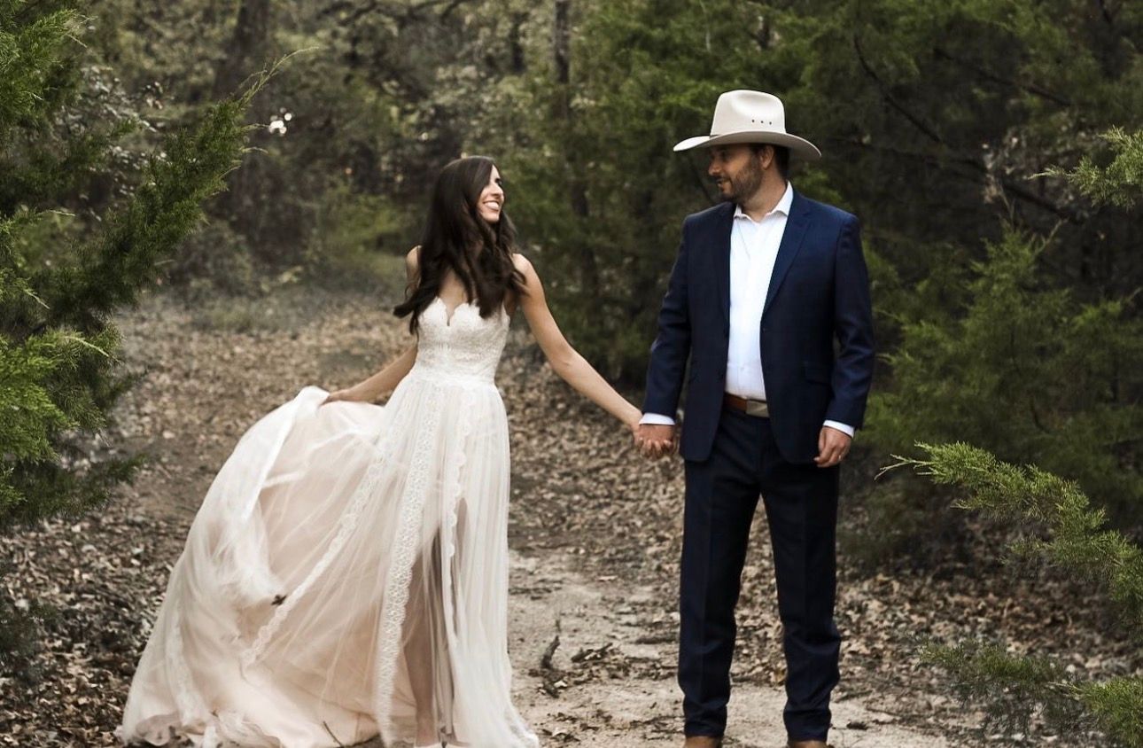 Bride in flowing champagne gown holds hands with groom in navy suit and cowboy hat in wooded outdoor wedding portrait.