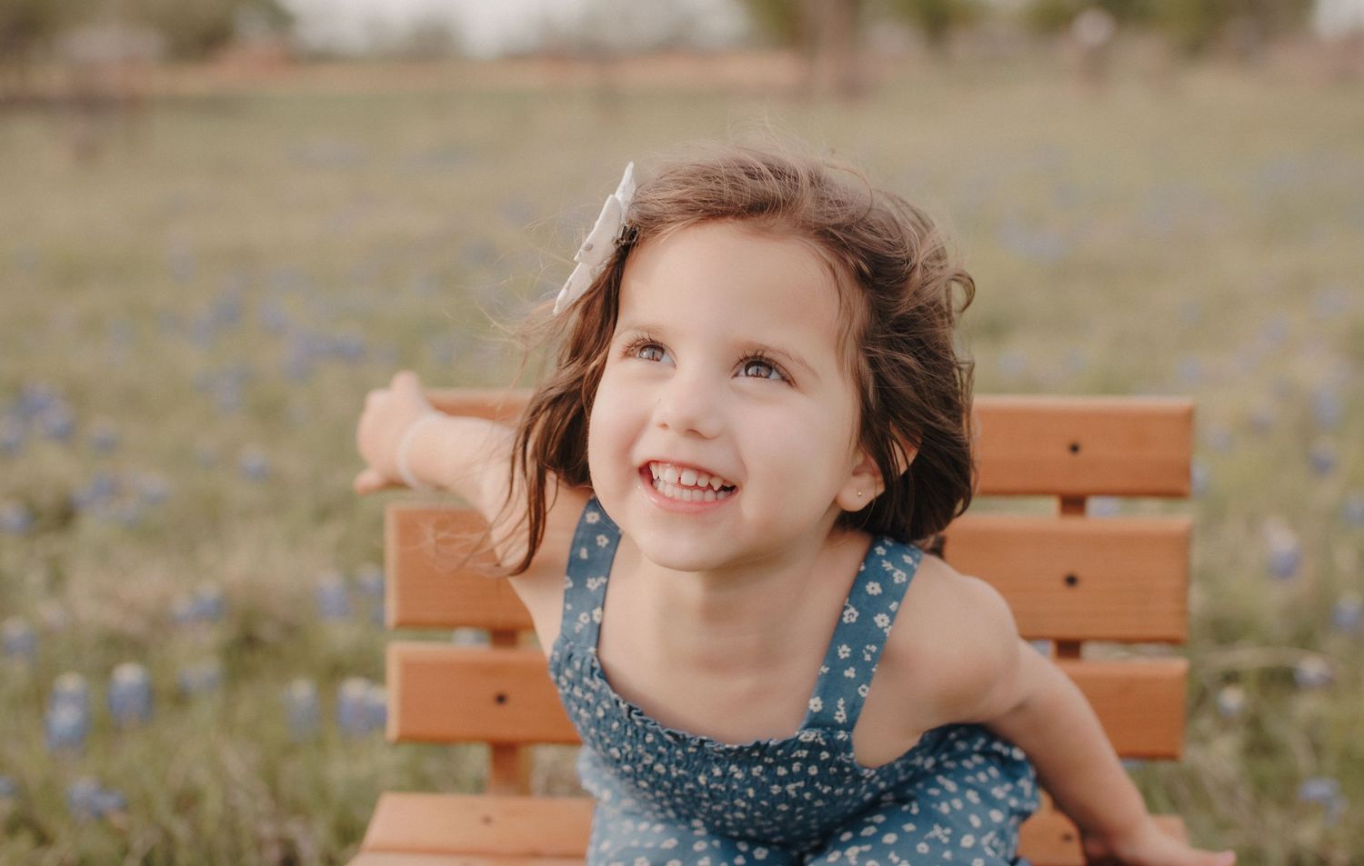 Cheerful young girl with curly hair sitting on wooden bench in field of blue wildflowers.