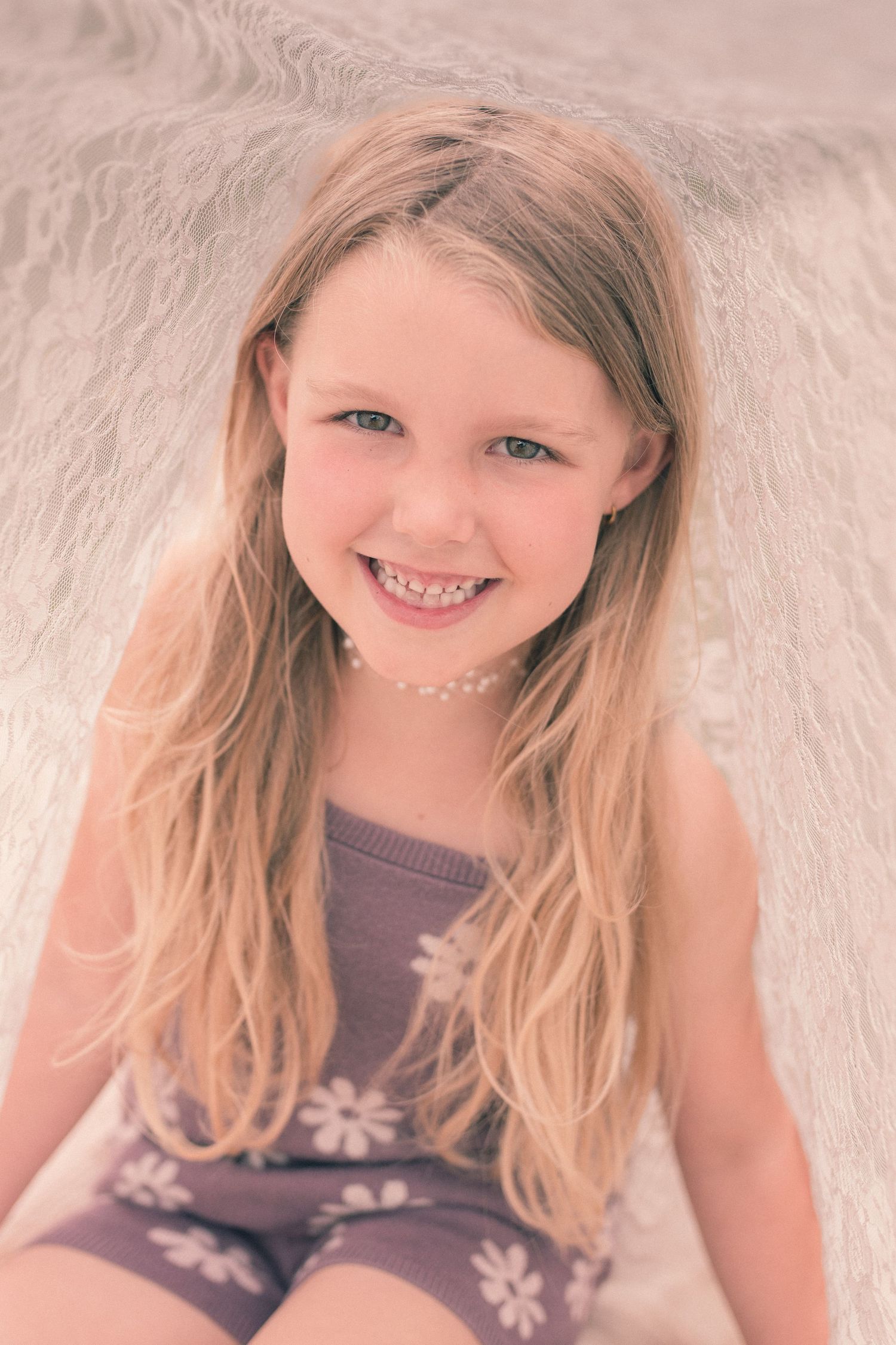 Smiling young girl with long blonde hair wearing a purple floral outfit, sitting against a soft white background.
