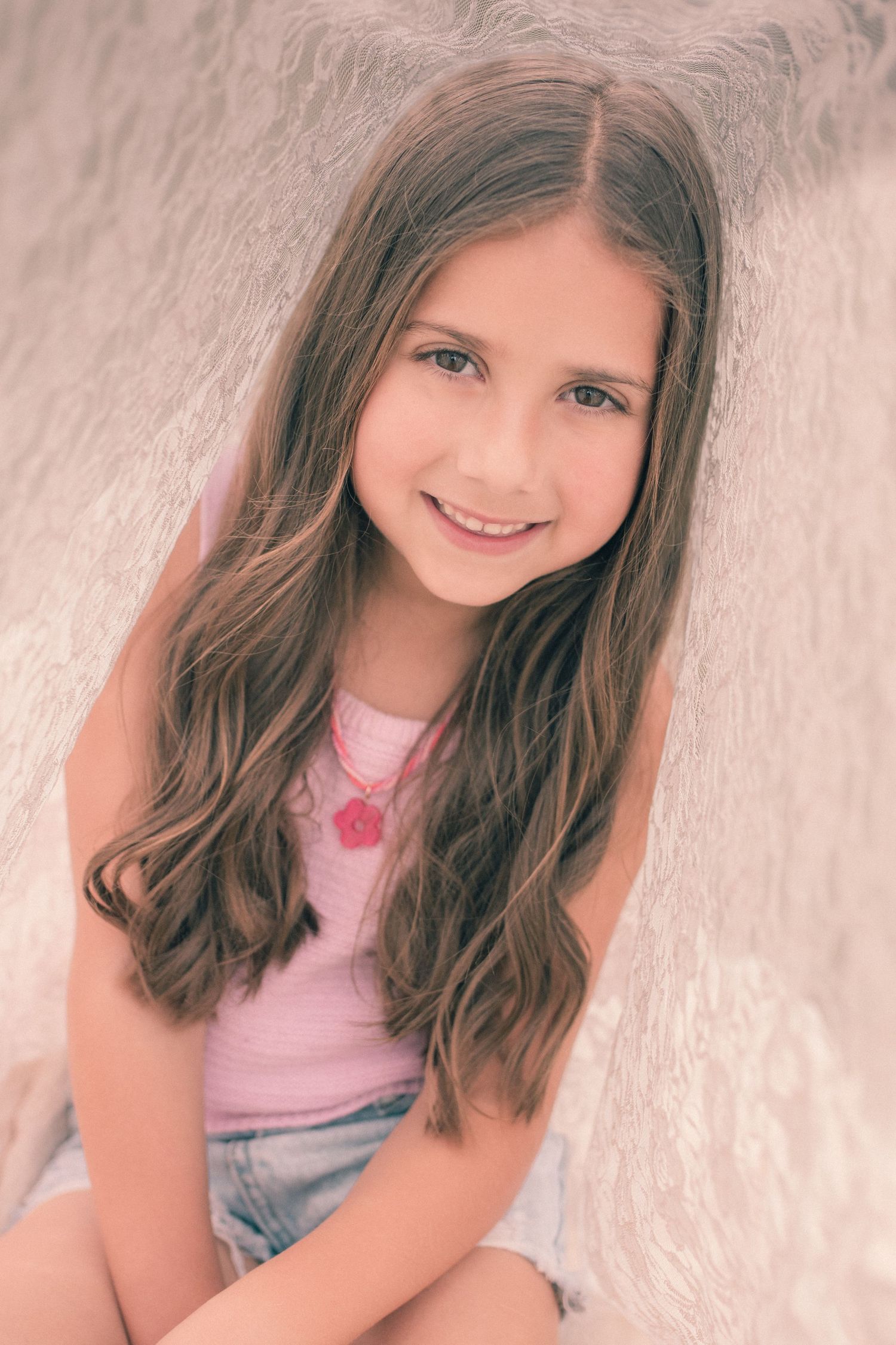 Smiling young girl with long brown hair wearing a pink top and red necklace, posed against a soft white background.