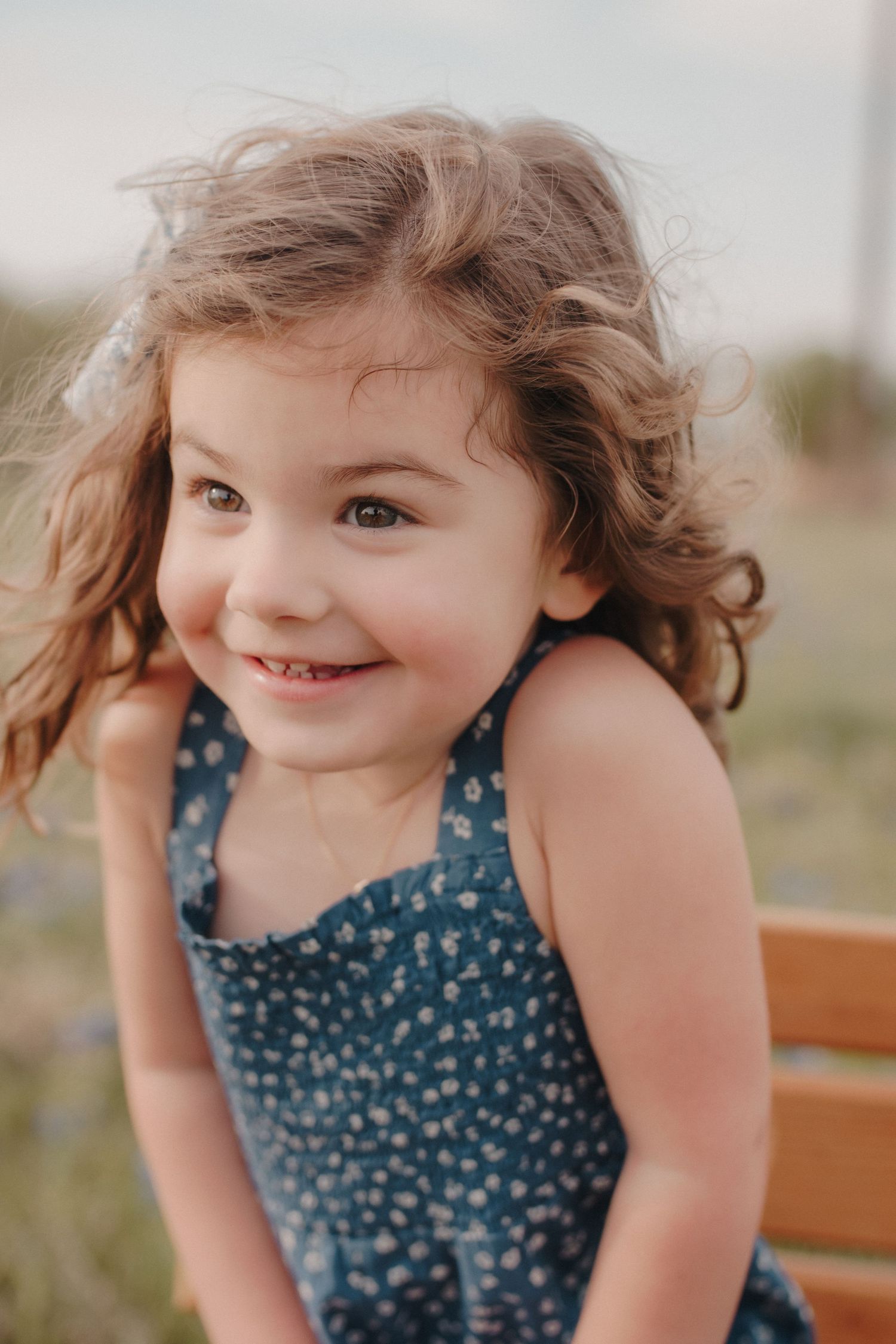 Smiling little girl with curly brown hair wearing blue floral dress outdoors at sunset.