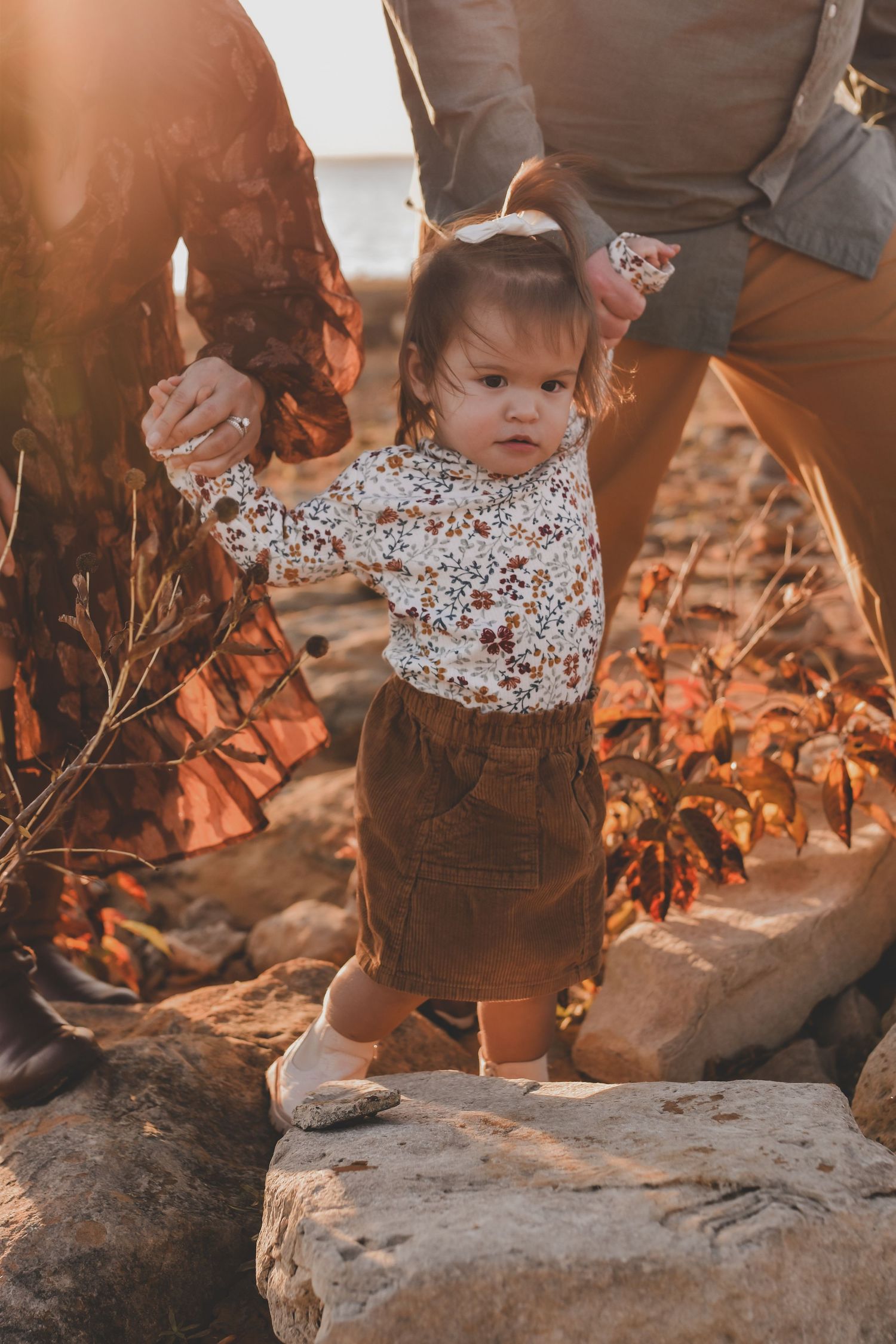 Toddler girl in floral top holds parents hands while standing on rocks during golden hour family photography session.
