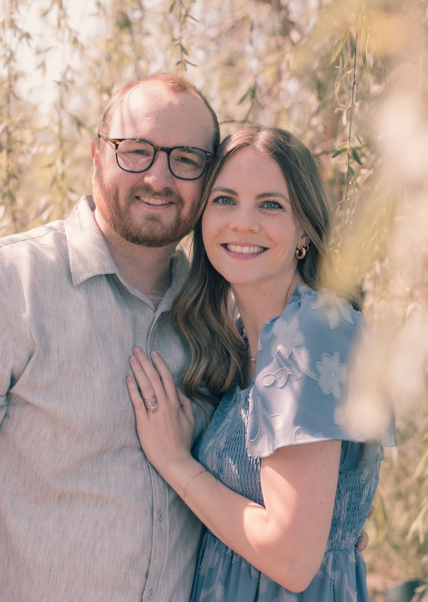 Smiling couple posing outdoors under willow tree branches, woman in blue dress, man in glasses and linen shirt.