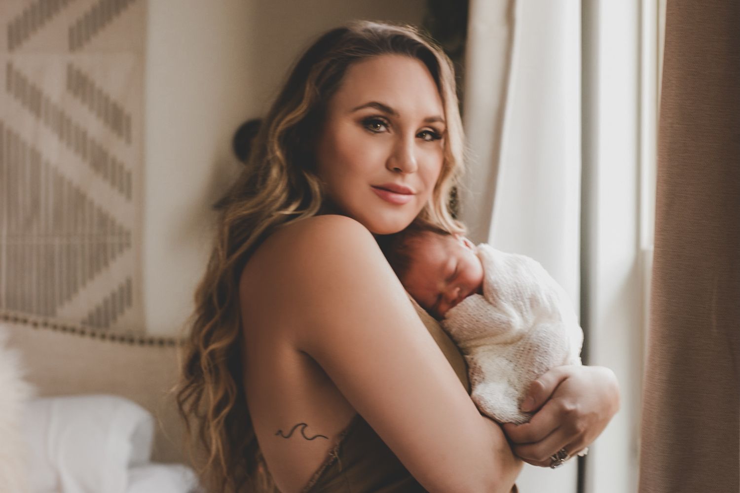New mother tenderly holding her sleeping newborn baby near a bright window indoors.