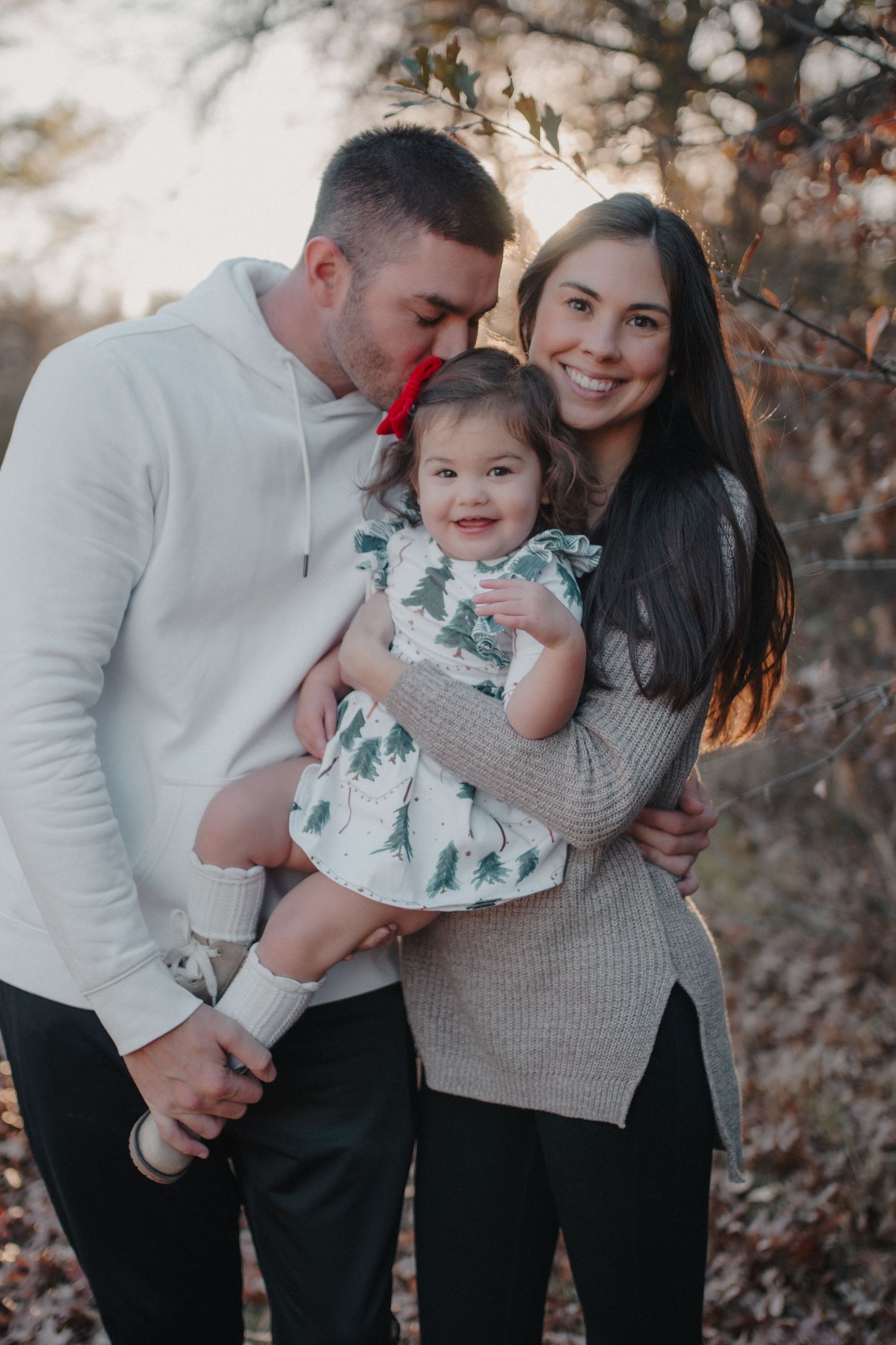 Happy family portrait with parents kissing toddler daughter outdoors in autumn setting.