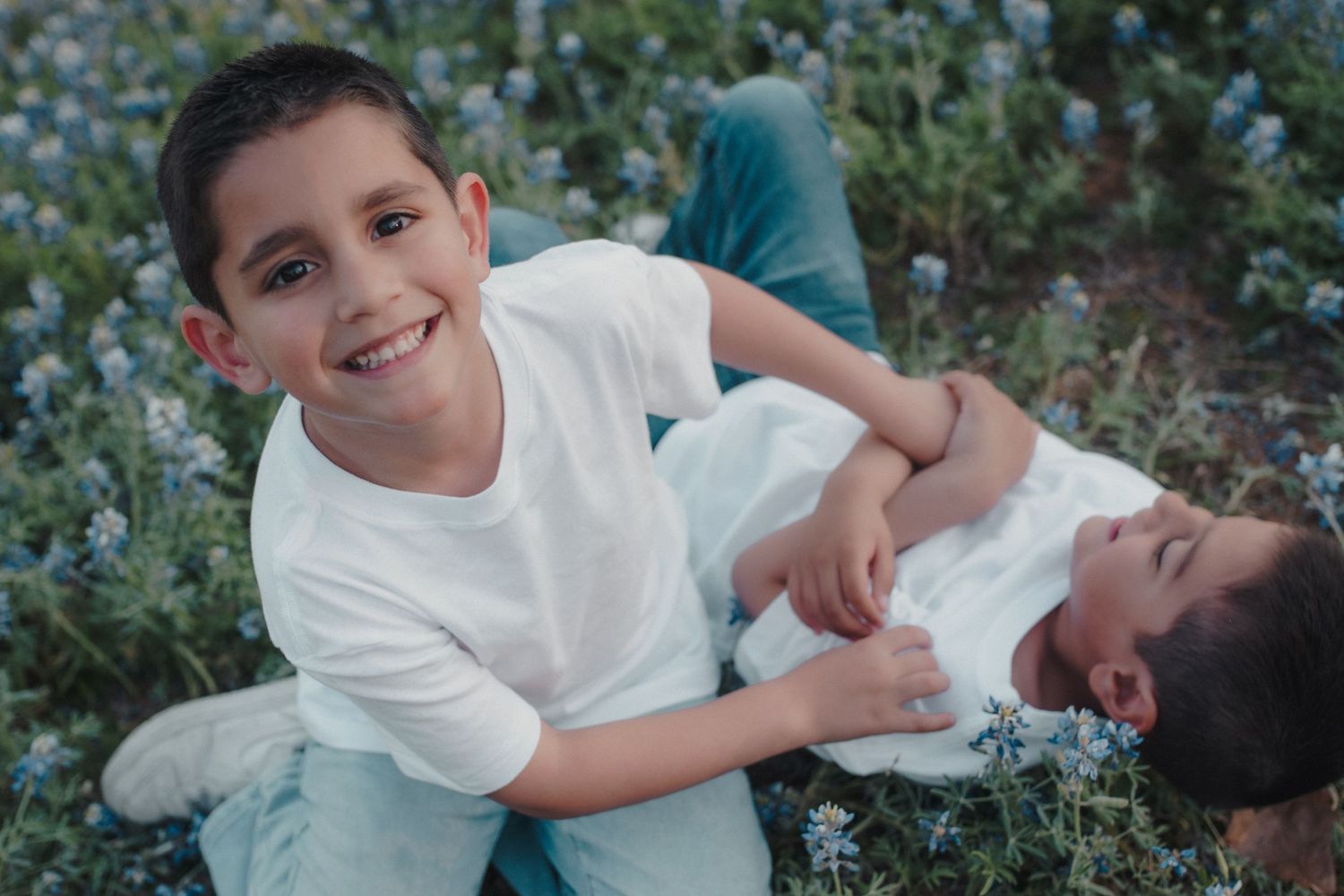 Two smiling boys in white shirts playing together in a field of blue wildflowers.