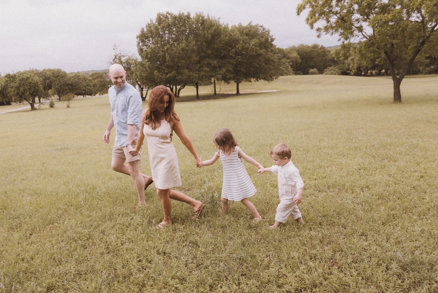 Family of four holding hands and walking through a lush green park on a warm sunny day.