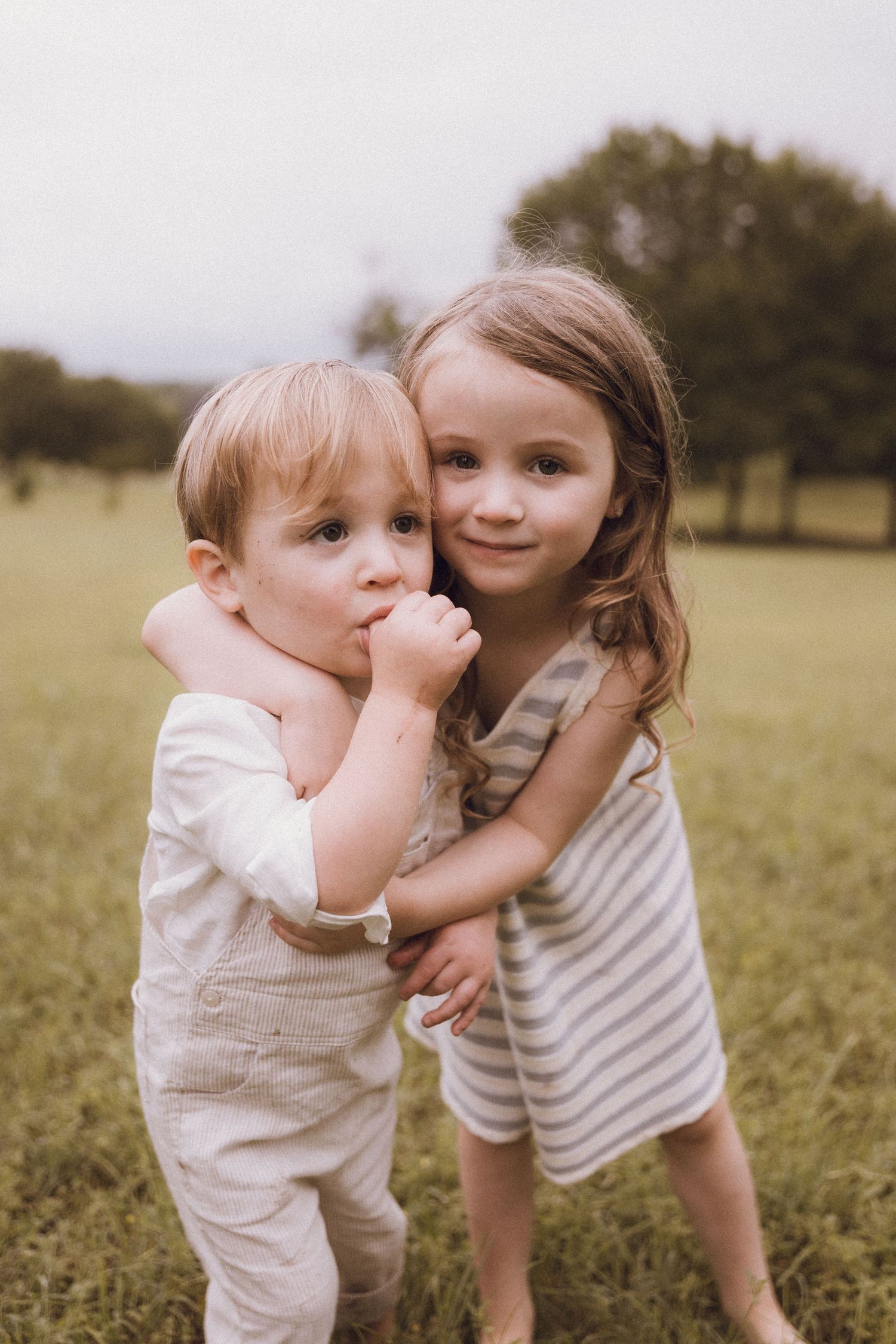 Two young siblings share a sweet hug in a grassy field, with an older girl embracing a toddler boy outdoors.