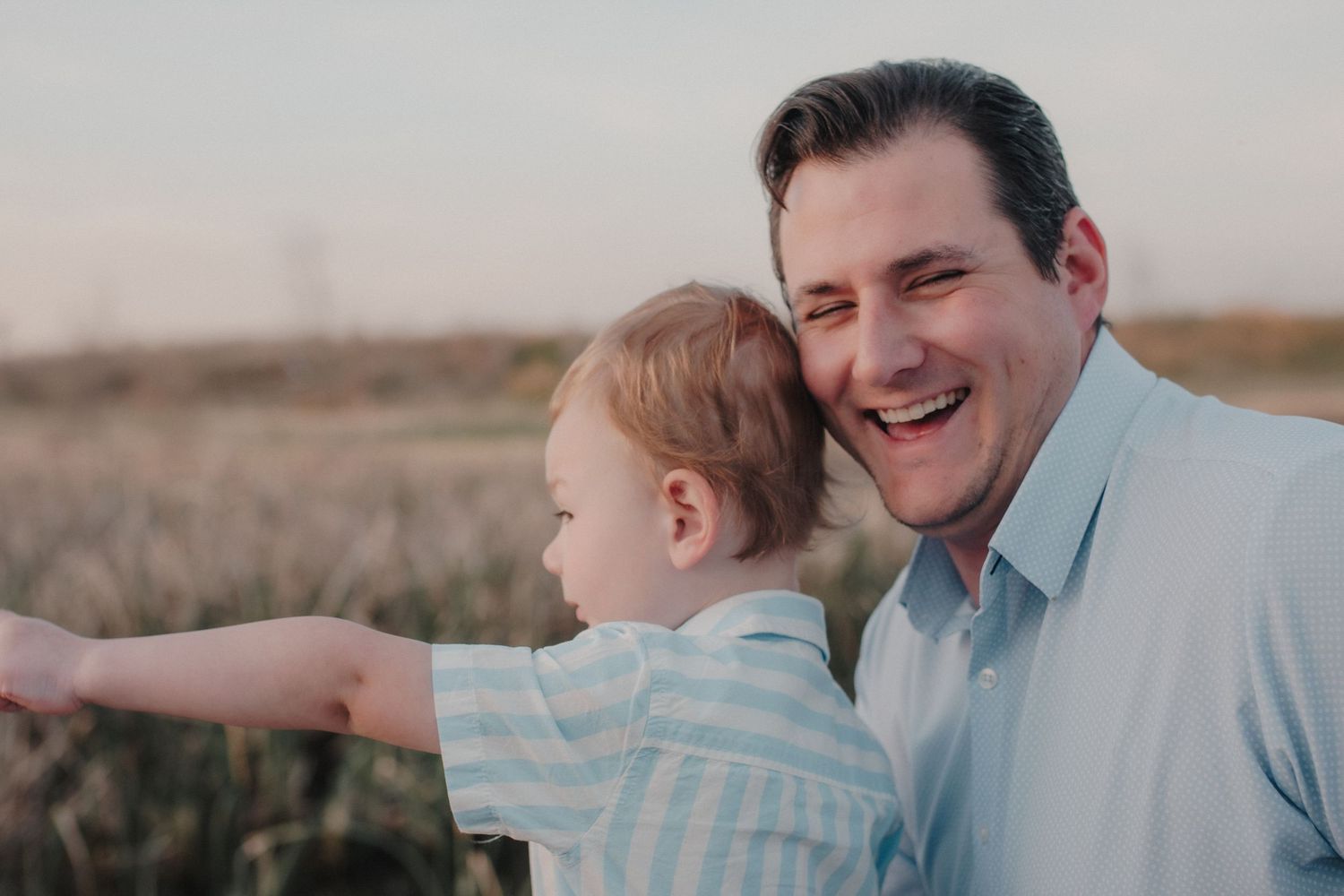 Laughing father holding happy toddler son in a sunny golden field.