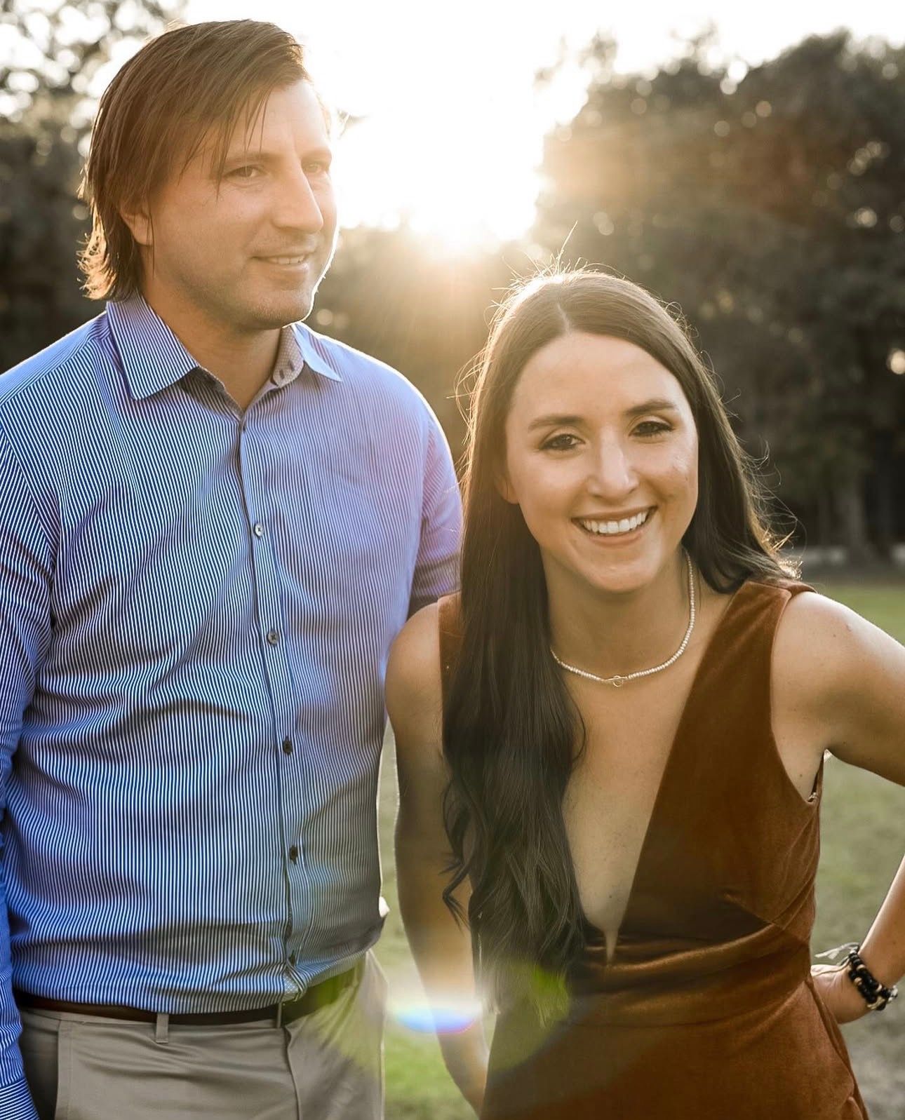 Smiling couple posing together outdoors with golden sunset light glowing through trees behind them.
