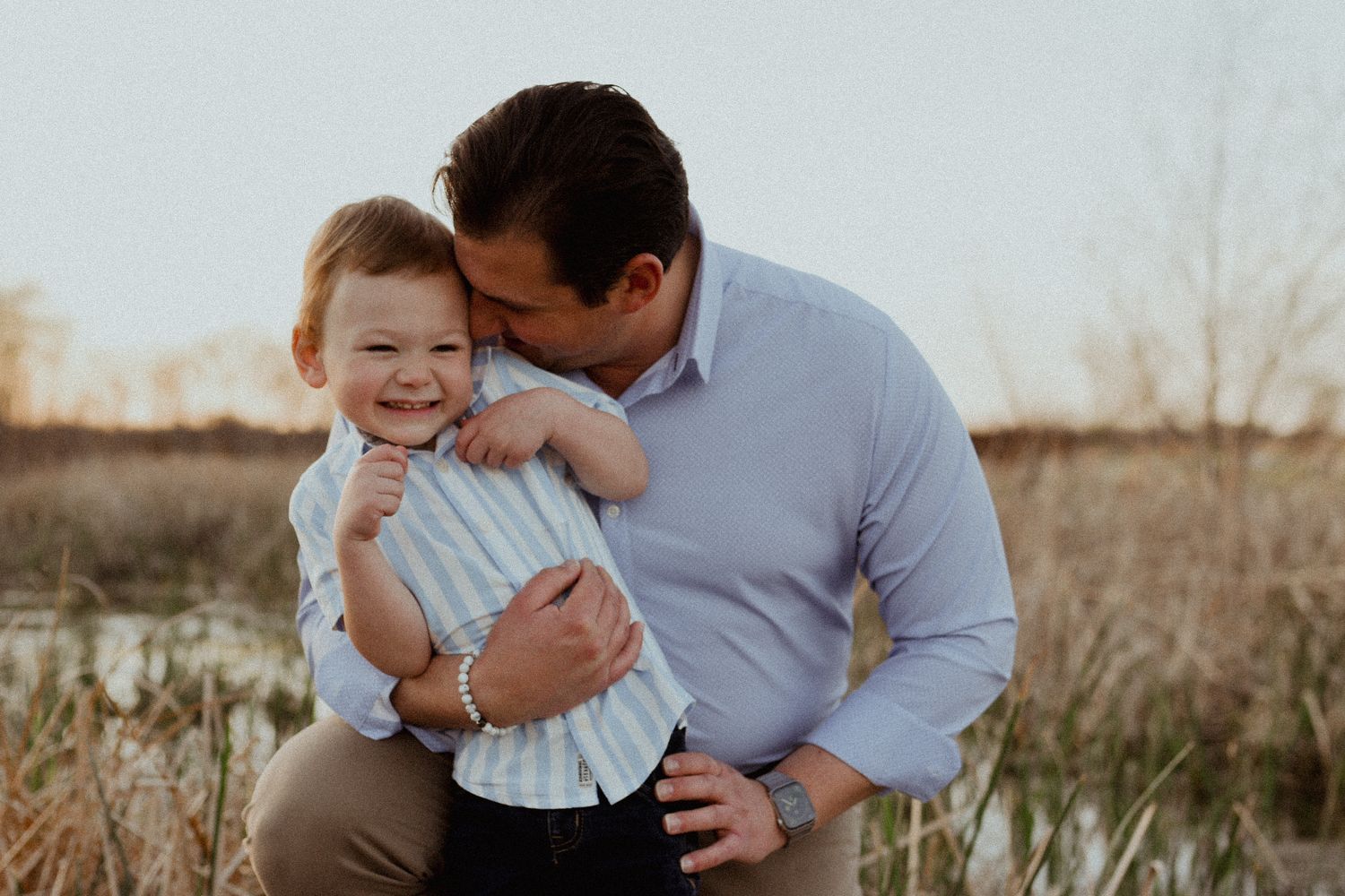 A father in a blue shirt kneels outdoors and kisses his laughing toddler son on the cheek.