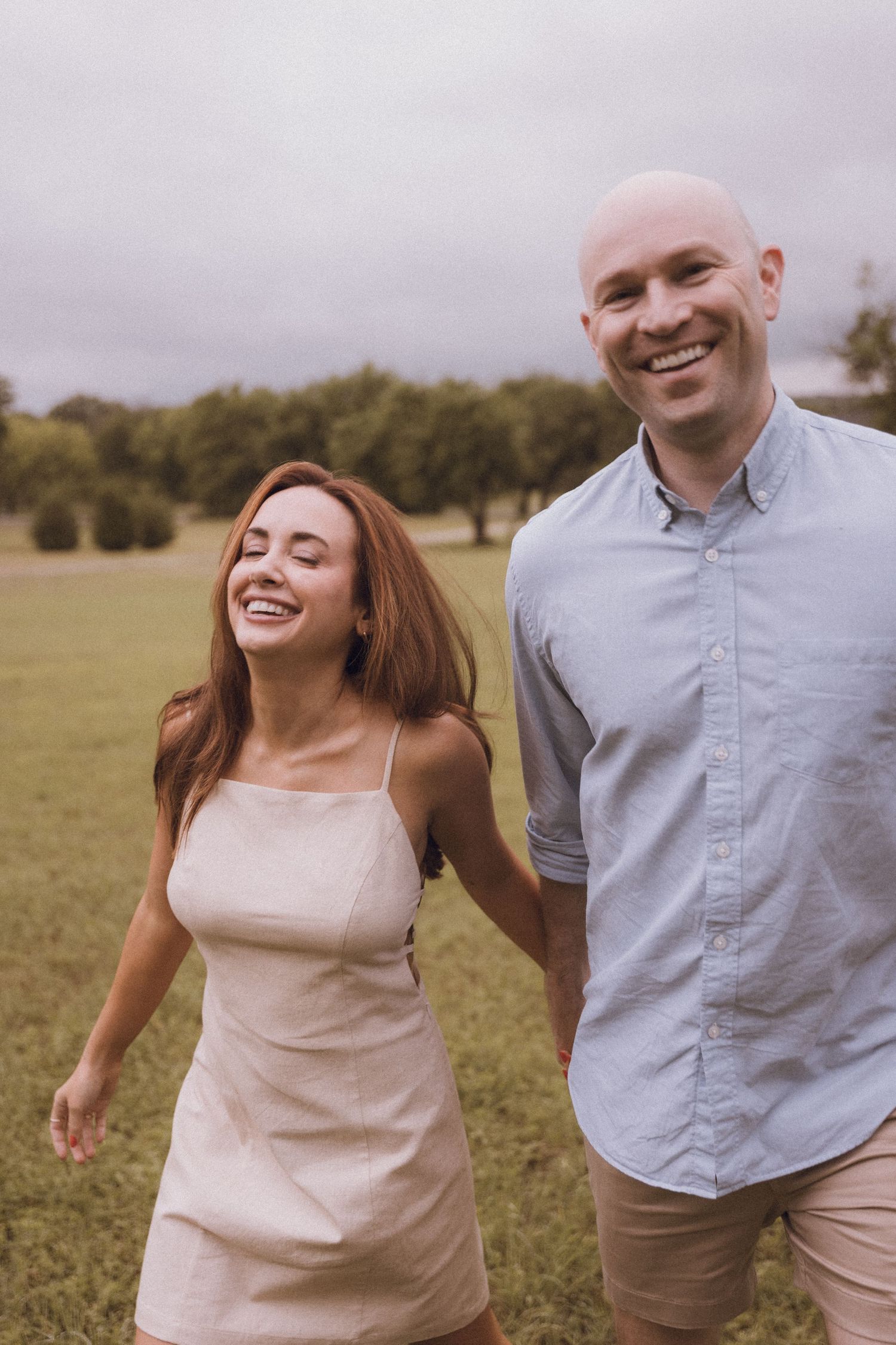 Happy couple holding hands and laughing while walking through a green park on a cloudy day.