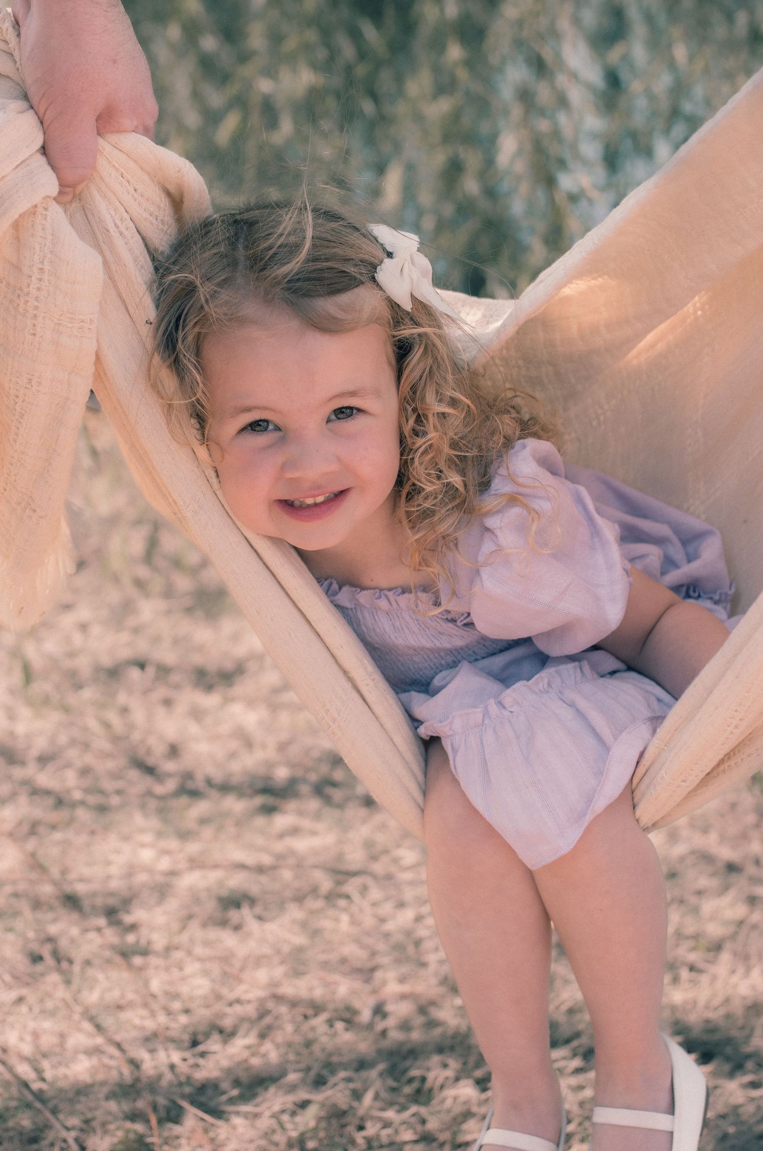 Smiling toddler girl with curly blonde hair in lavender dress sitting in a fabric swing outdoors.