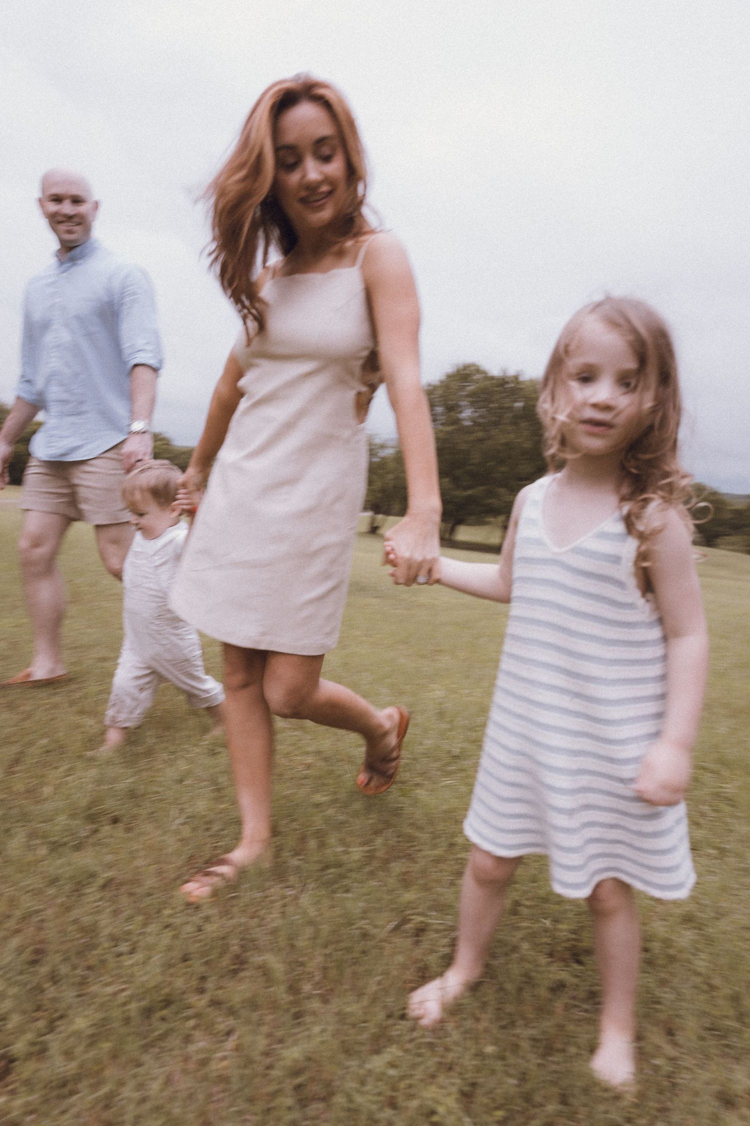 A happy family walks barefoot on green grass, with a woman in a beige dress holding a young girl's hand.