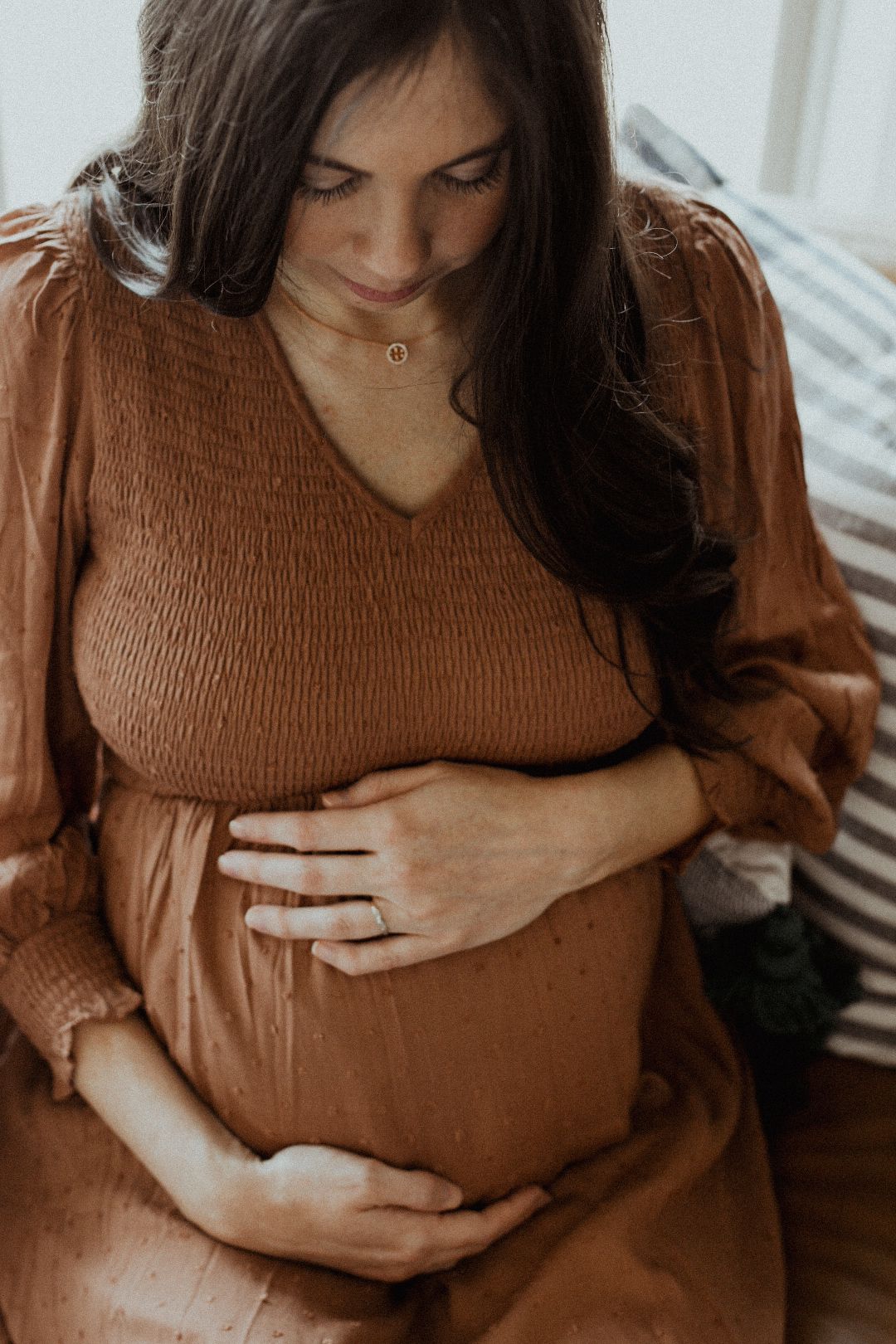 A pregnant woman in a rust-colored dress gently cradles her baby bump while looking down softly.